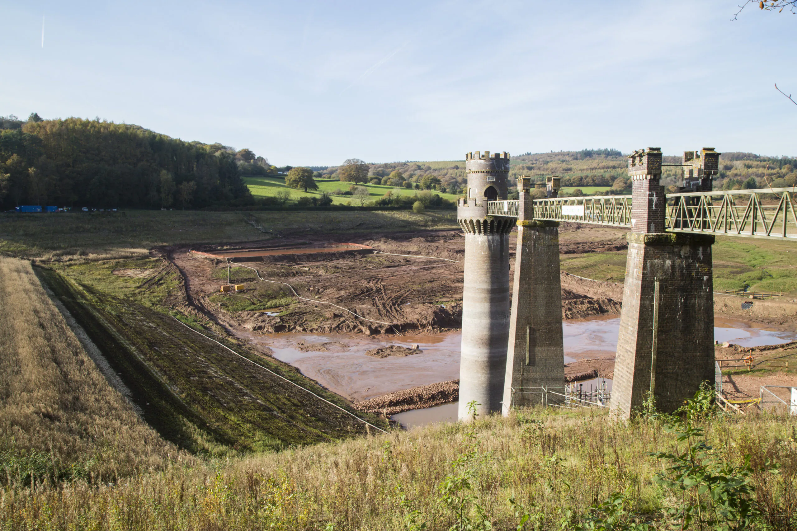 Old stone bridge with two towers over an excavated area, possibly part of a dam or reservoir project in a countryside setting.