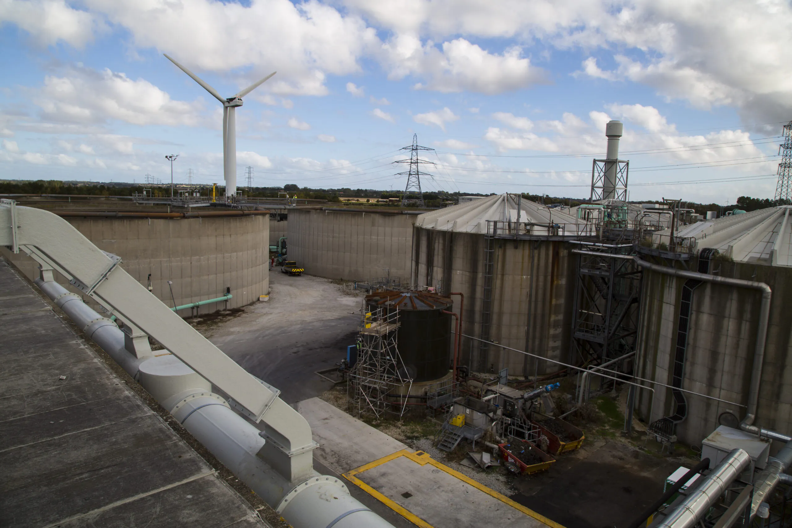 Industrial facility with large storage tanks, pipelines, wind turbines, and power lines under partly cloudy skies.