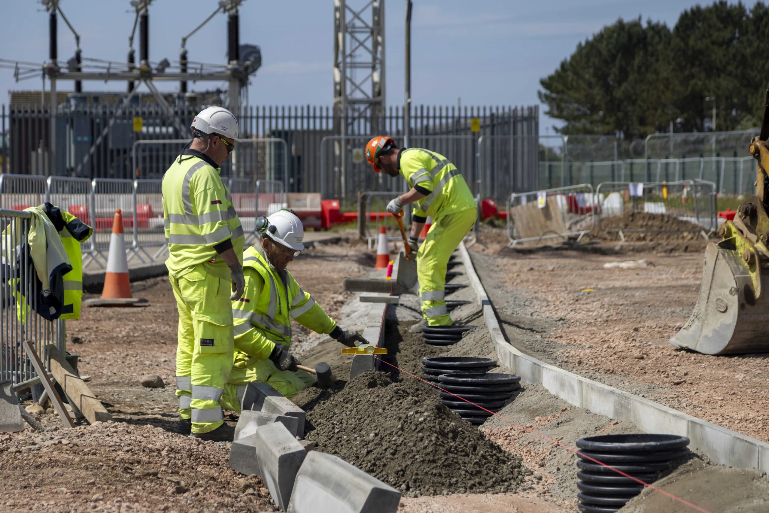 Three construction workers in high-visibility clothing and helmets work on-site; one measures the ground, another digs with a shovel, and the third observes. Safety cones and equipment are visible.