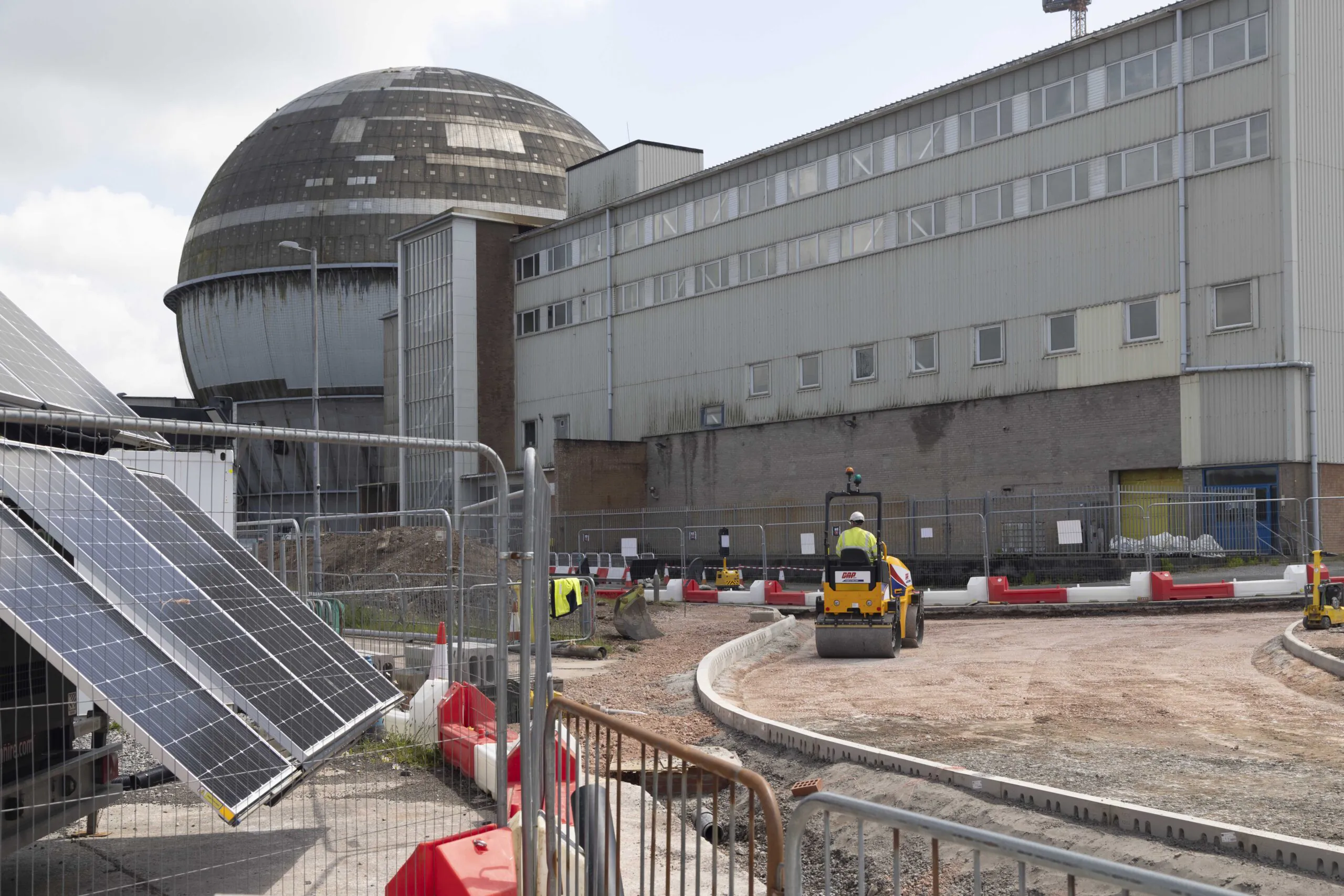 Construction workers operate machinery near an industrial building with a dome structure; solar panels, fencing, and barriers are visible in the foreground.