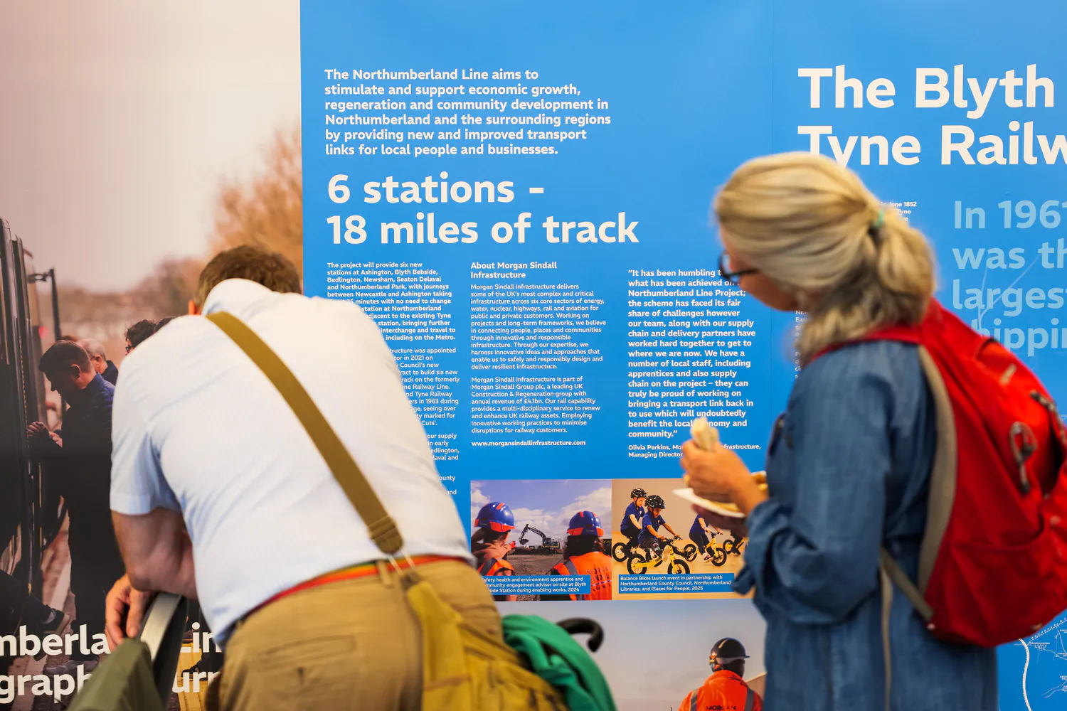 Two individuals reading a display board about the Northumberland Line project, which outlines plans for six stations and 18 miles of track to boost regional transport and economy.