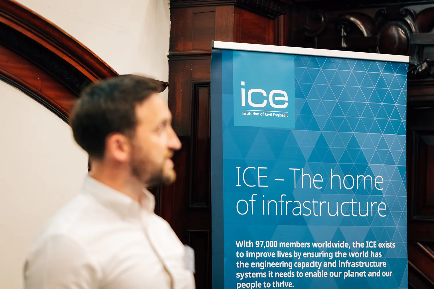 Person standing next to a banner reading "ICE - The home of infrastructure," describing the Institution of Civil Engineers' mission.