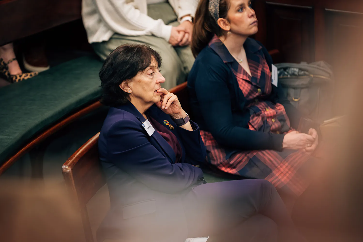 Attendees listening attentively during an indoor event; one woman dressed formally in the foreground, another in a plaid dress.
