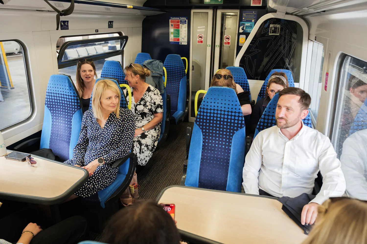 Inside a train carriage, people seated at tables and chairs engaged in conversation or observing surroundings.