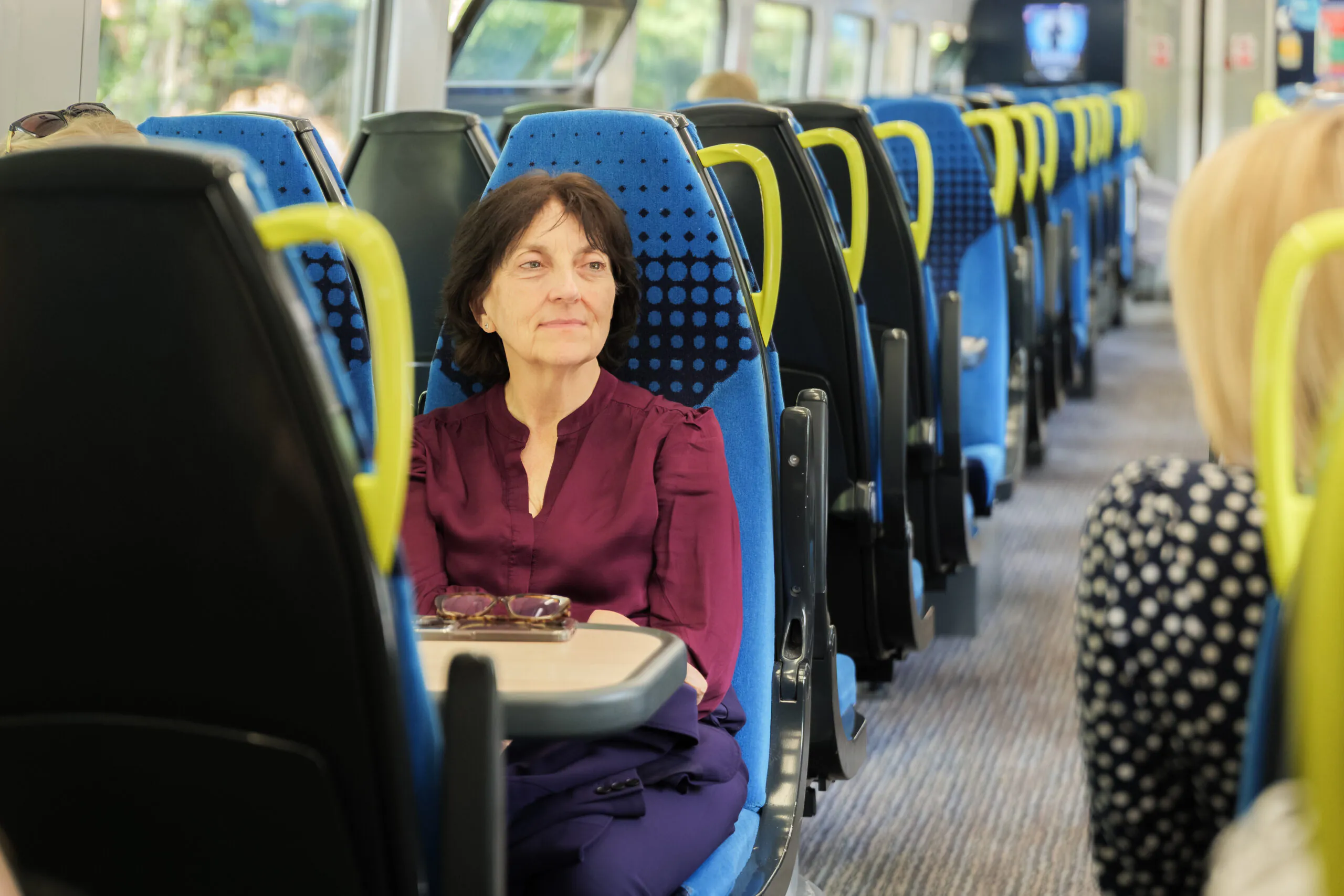 Person seated on a modern train with blue seats featuring black polka dots and yellow handles, looking out the window. Rows of similar seats extend into the background.