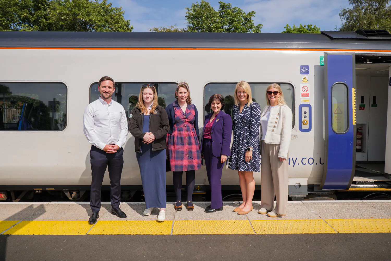 Six individuals posing for a group photo on a train platform in front of a white and blue train. The platform includes yellow tactile paving for safety.