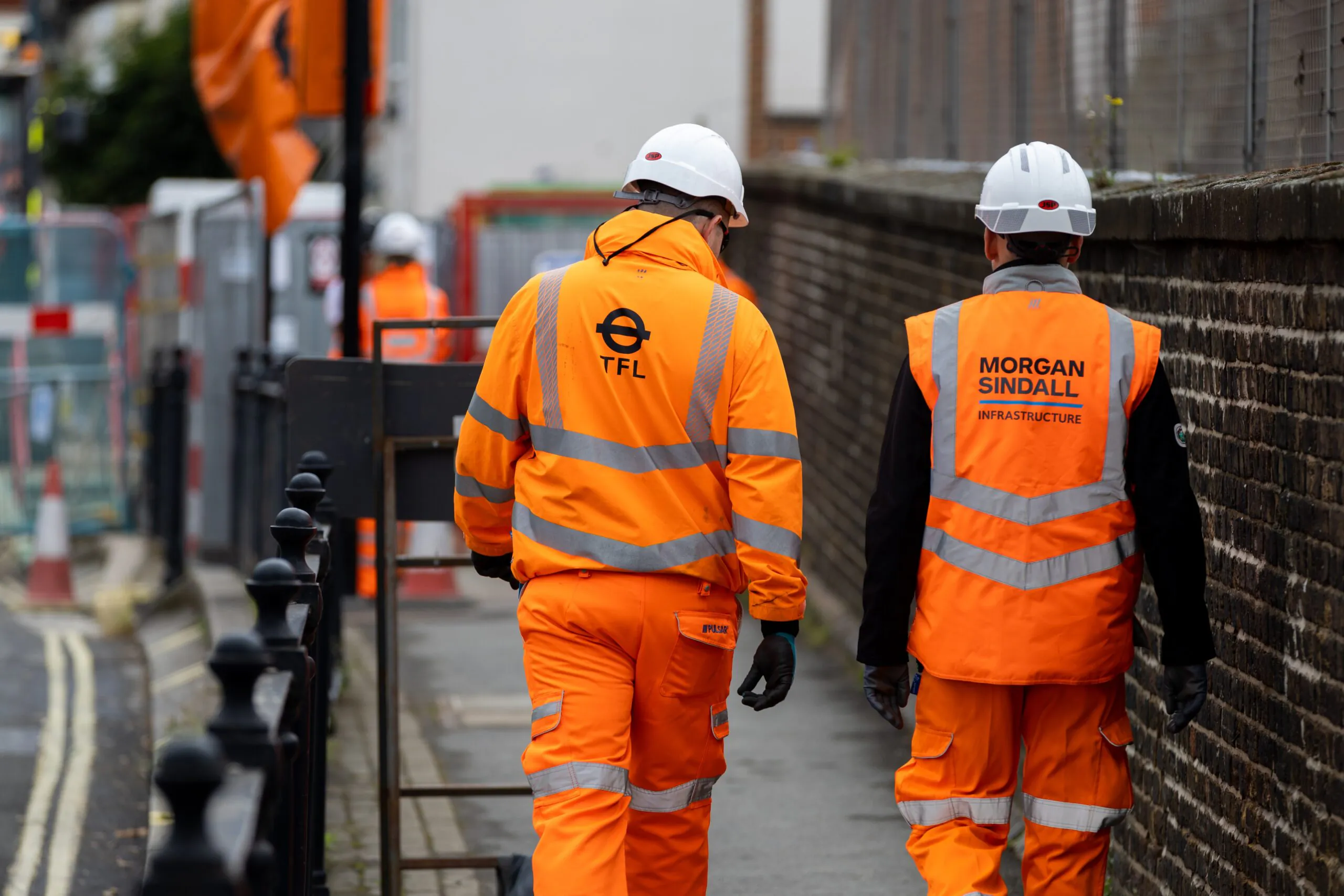 Two people in orange high-visibility jackets walk down a street; one jacket reads "TFL" and the other "Morgan Sindall Infrastructure."