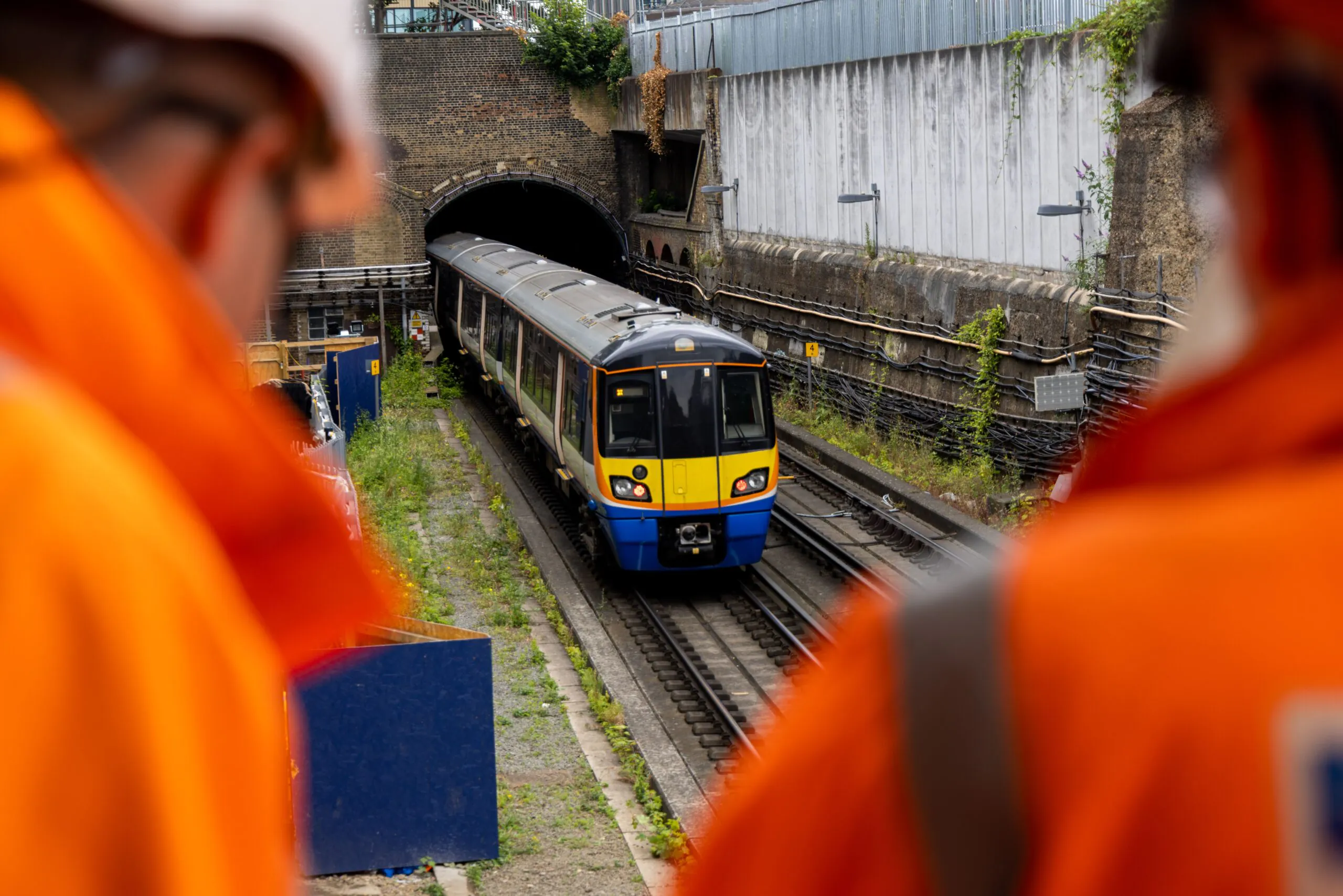 Train entering or exiting a tunnel viewed from above, with two individuals in orange safety gear standing in the foreground.