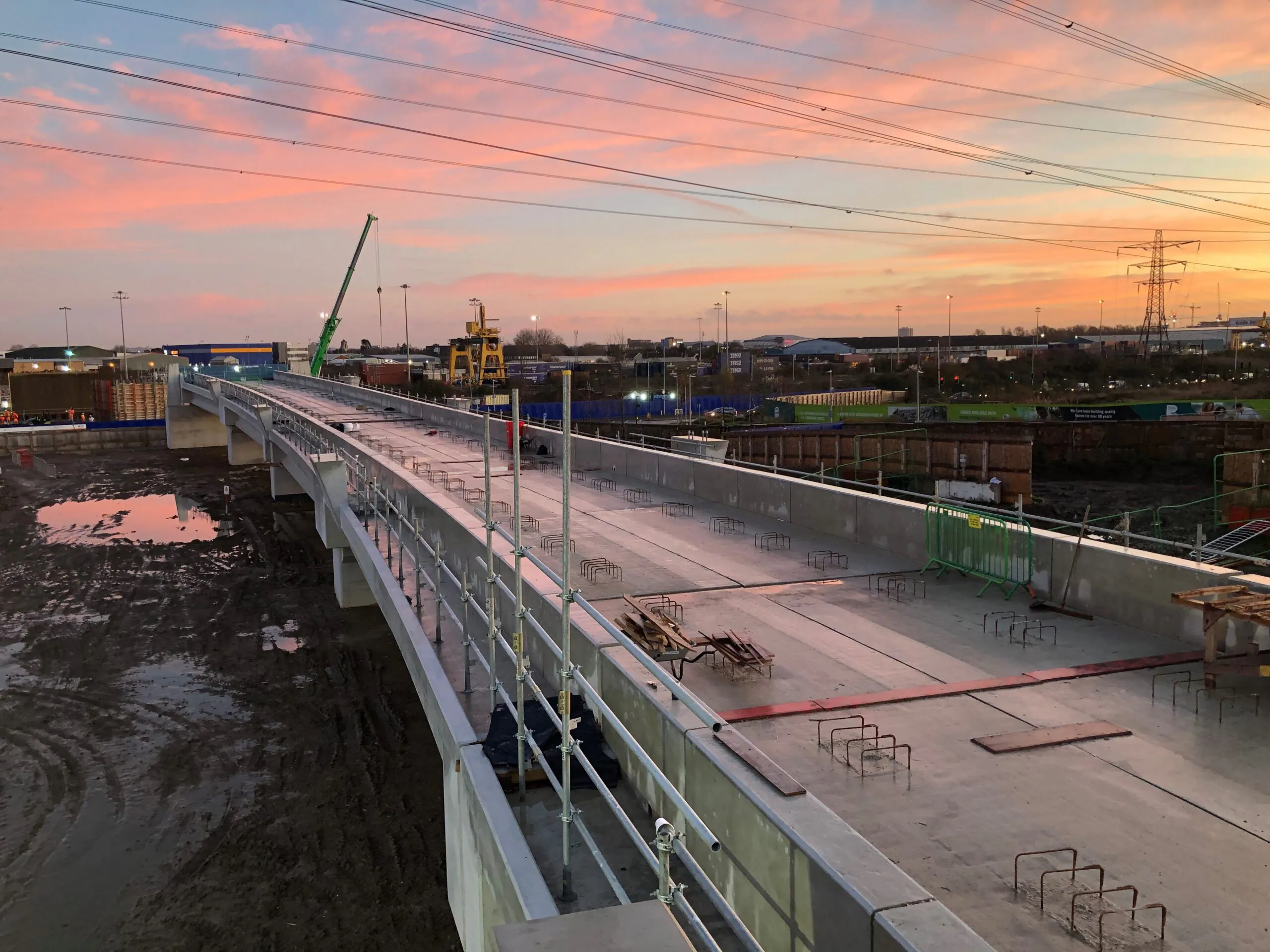 Photograph of a partially built bridge at sunset, featuring concrete sections, metal railings, cranes, and power lines silhouetted against the sky.