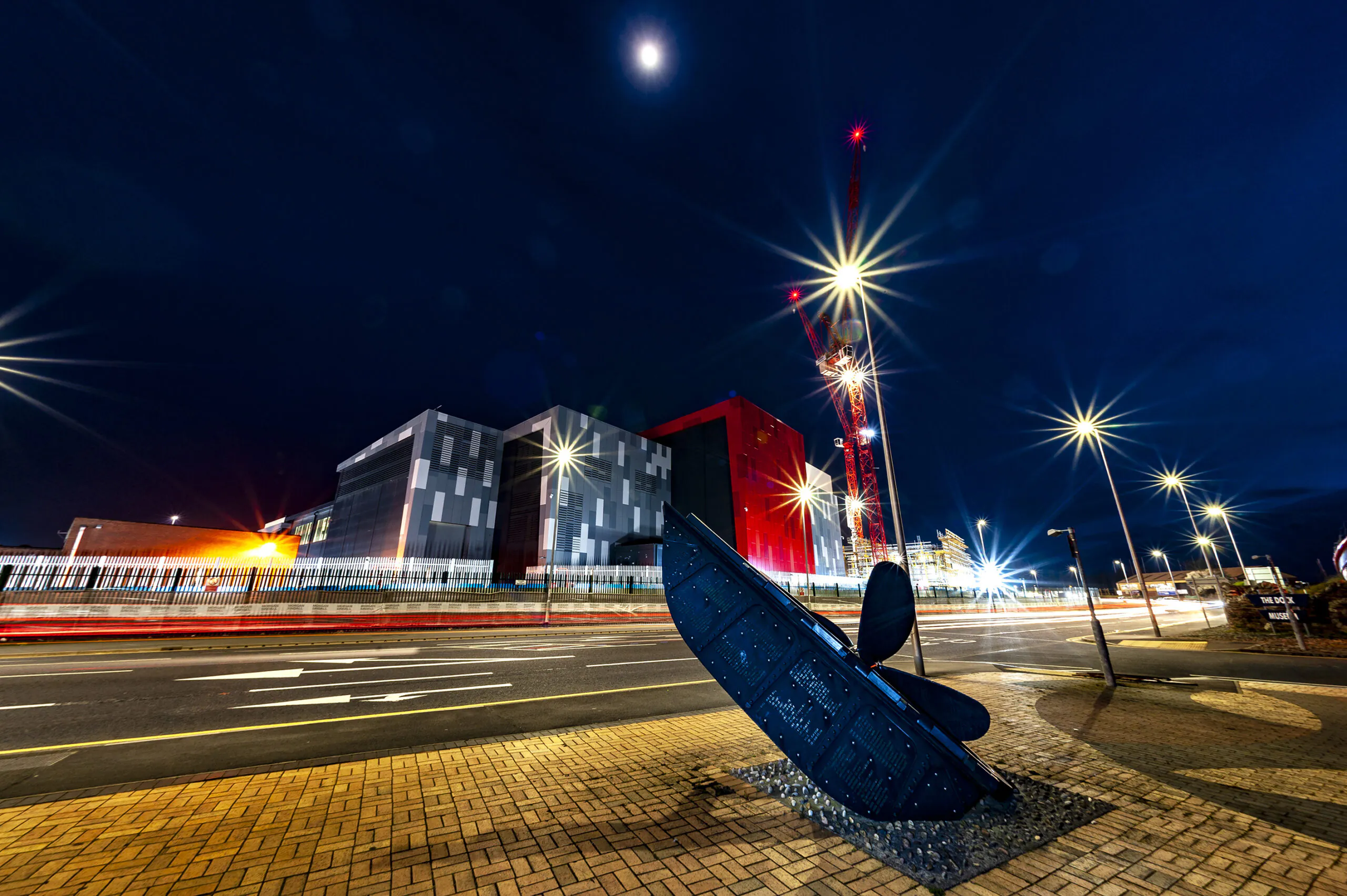 Nighttime view of an illuminated industrial building complex with light trails from passing vehicles and a large propeller sculpture in the foreground under moonlight.