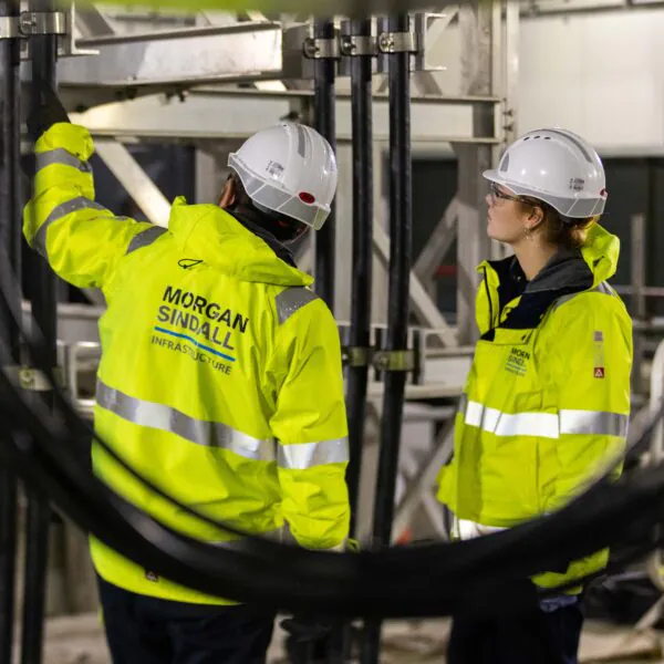 Two workers in high-visibility jackets and hard hats labeled “Morgan Sindall Infrastructure” inspect equipment in an industrial setting with cables and machinery.