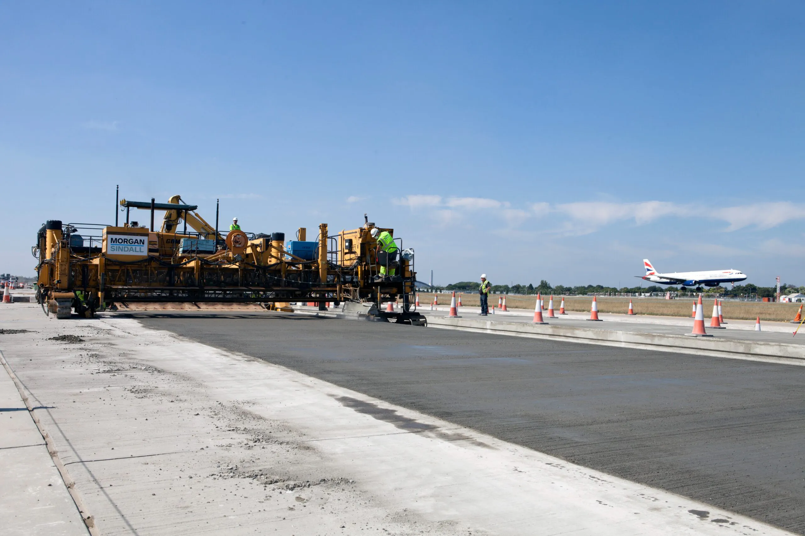 Large construction machine labeled "Morgan Sindall" paving concrete at an airport with workers in high-visibility clothing; airplane taking off in the background.
