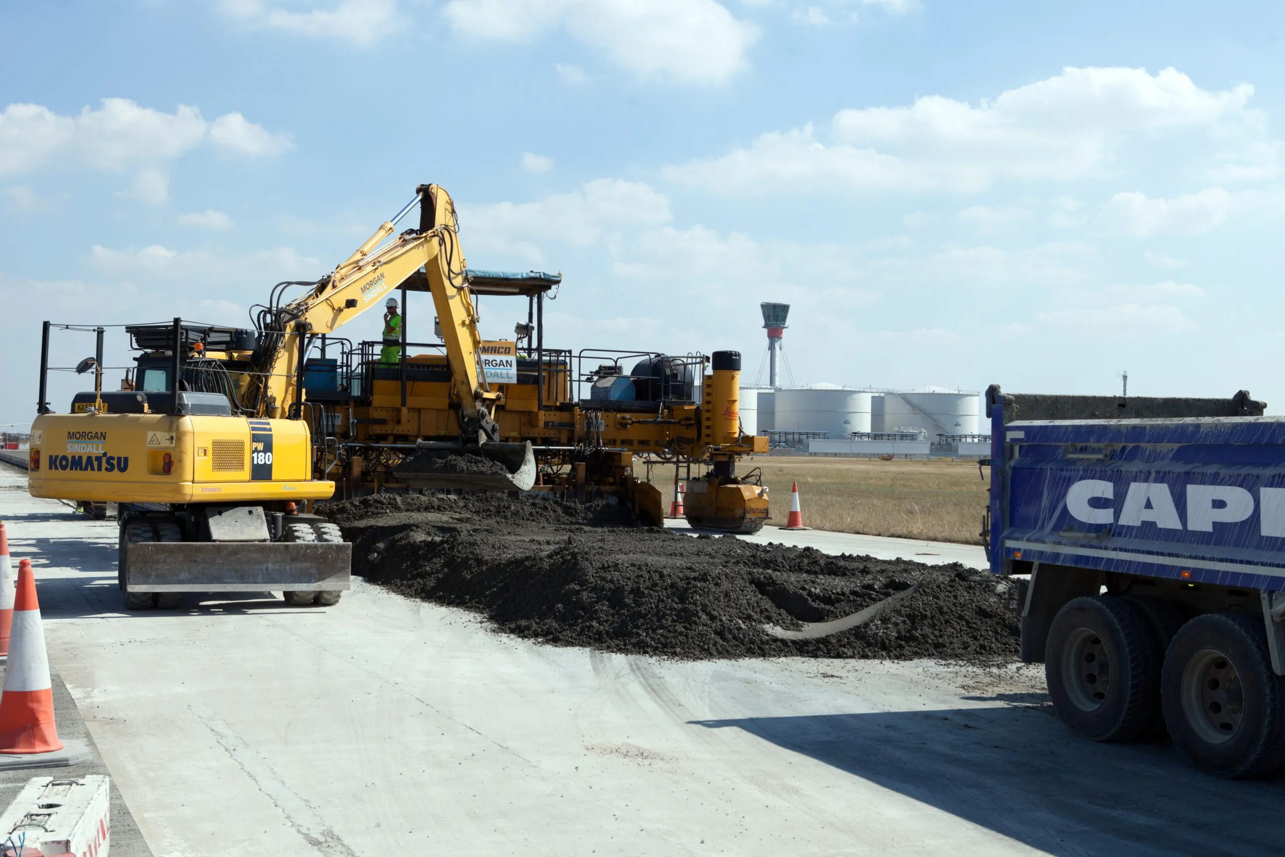 Excavators and trucks lay materials for an airport runway or taxiway; workers are present around the machinery.
