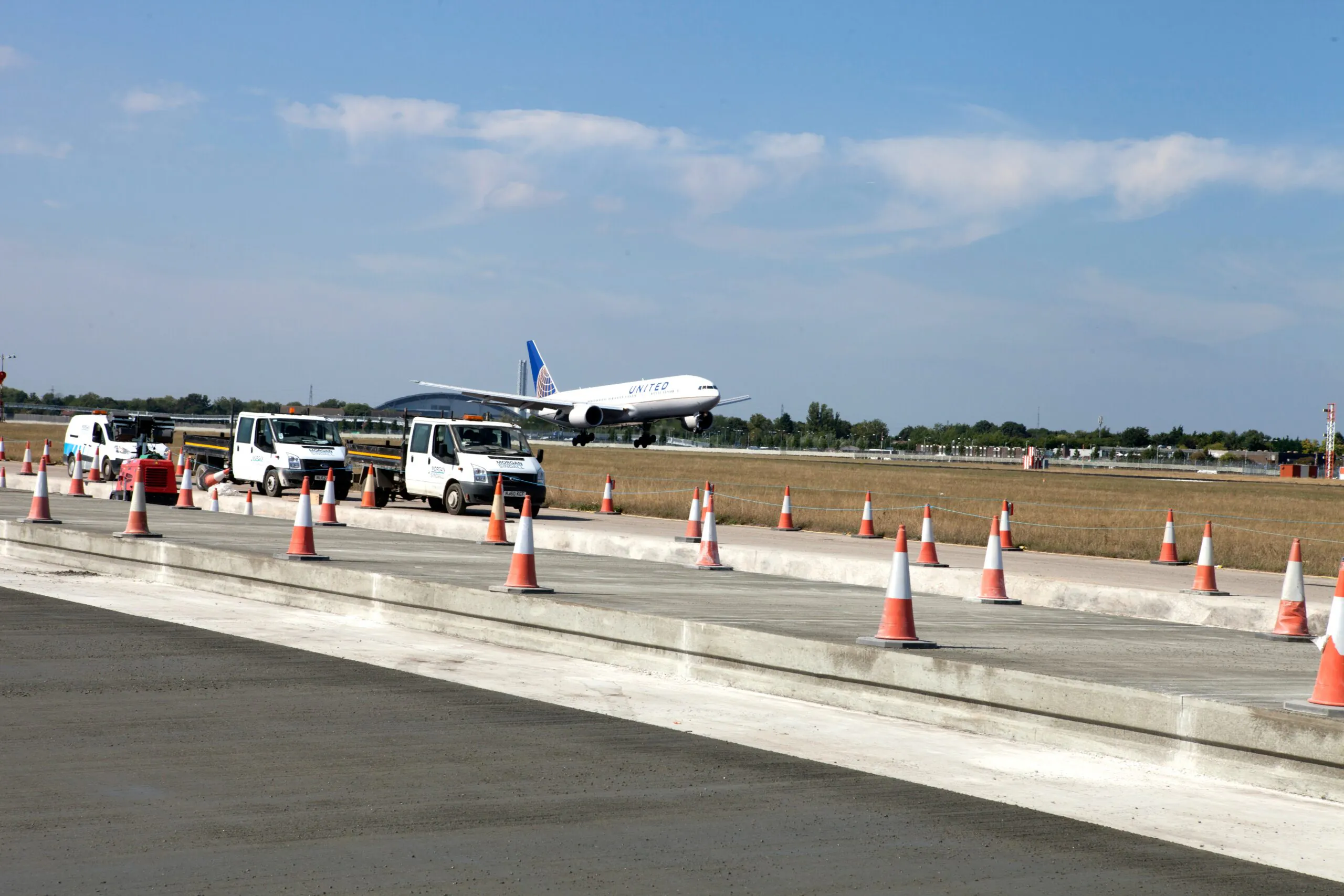 White utility vehicles parked along a newly paved airport runway section marked by traffic cones; a United airplane is landing in the background.