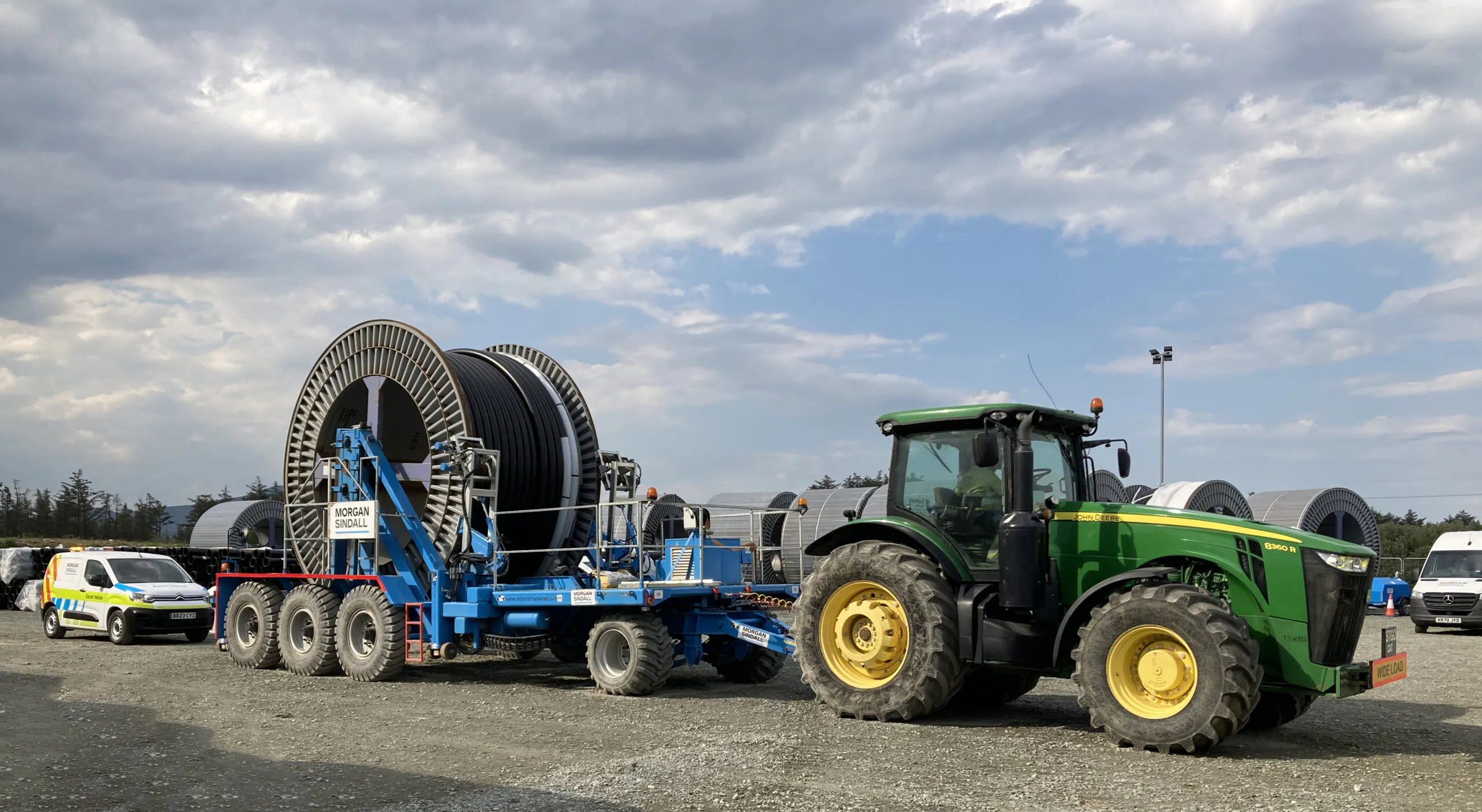 Green tractor equipped with winching gear parked on a dirt path in a rural setting under a partly cloudy sky.