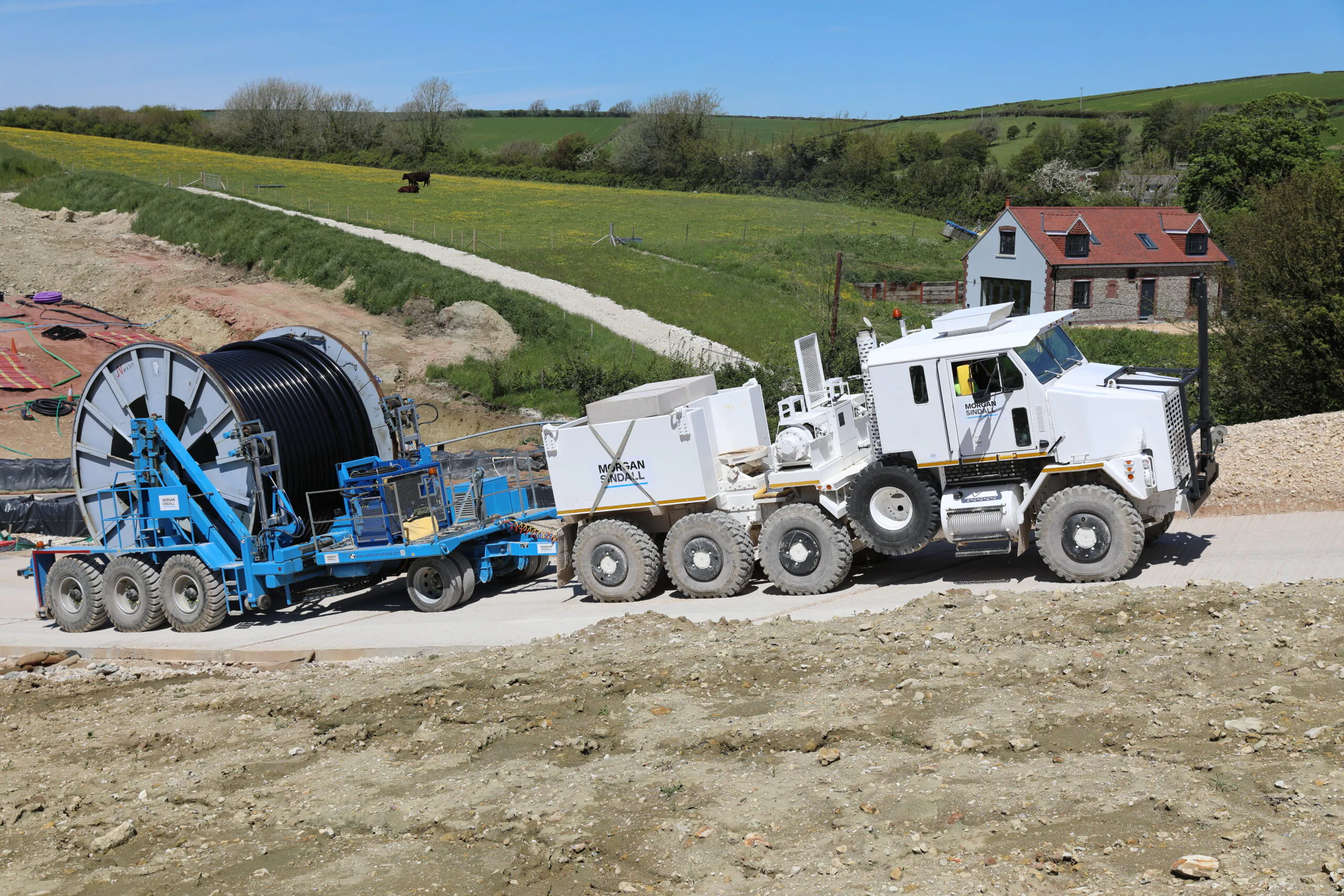 Massive off-road vehicle towing cable reels on trailers marked "Morgan Sindall," traveling through hilly countryside.