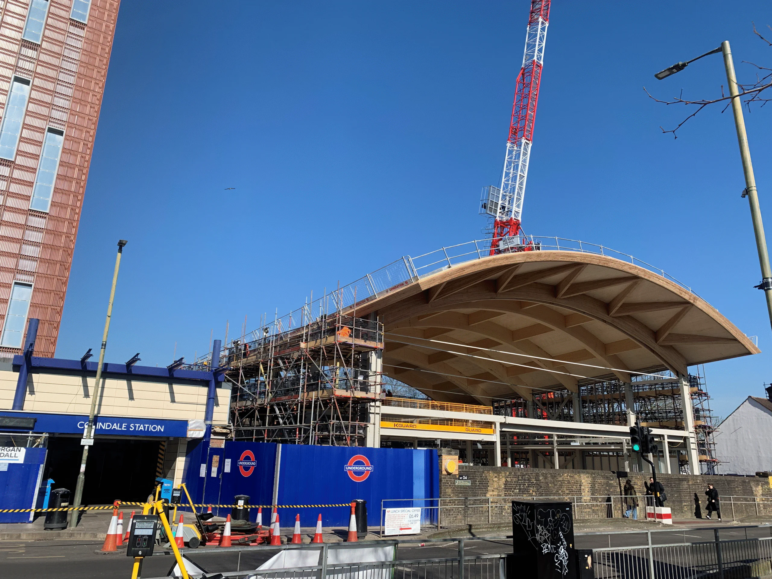 Daytime view of Colindale Station under construction with a large wooden arched roof and surrounding scaffolding; pedestrians nearby.
