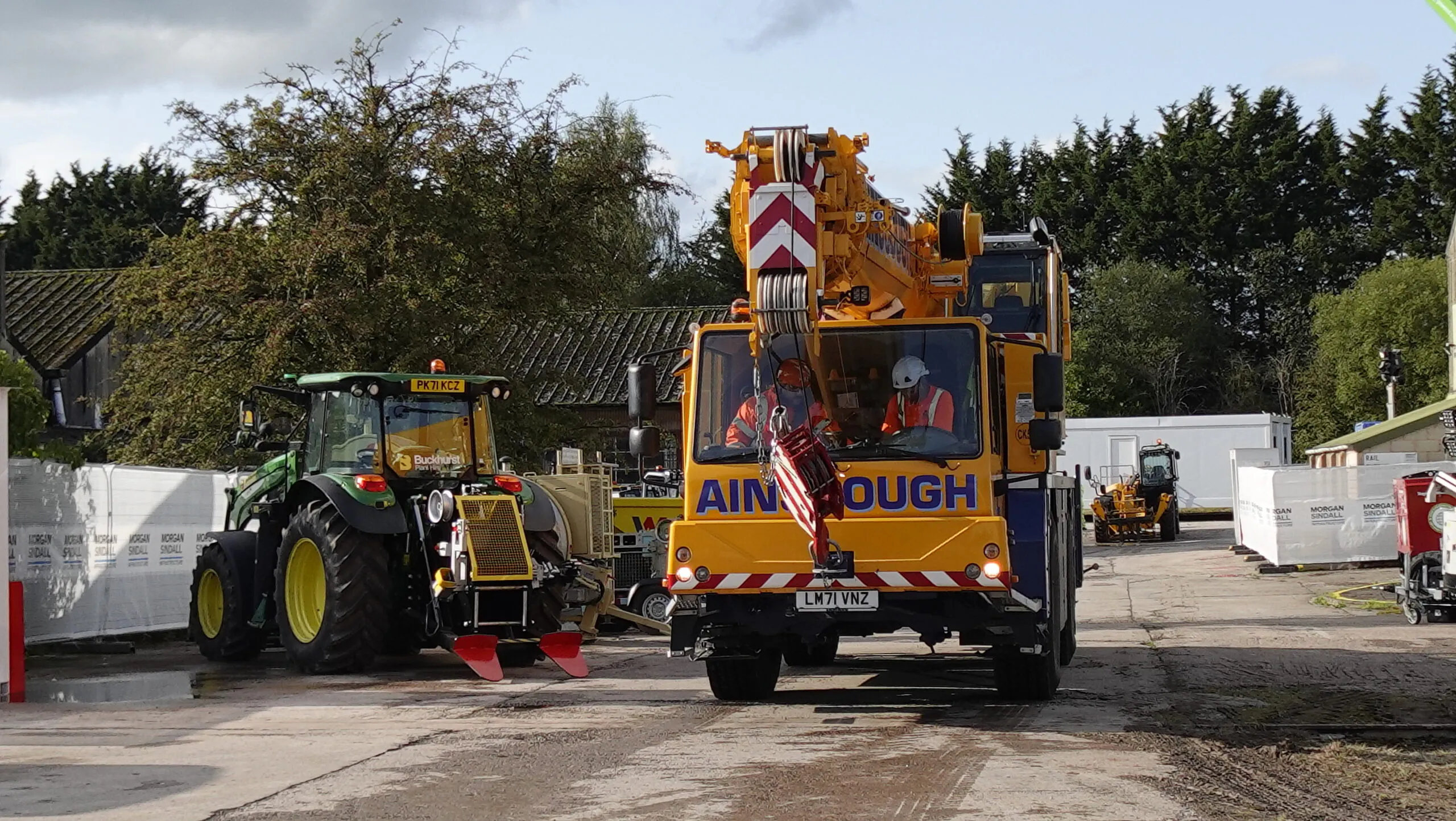 Construction vehicles including a tractor and crane moving through a site, with workers inside the vehicles.