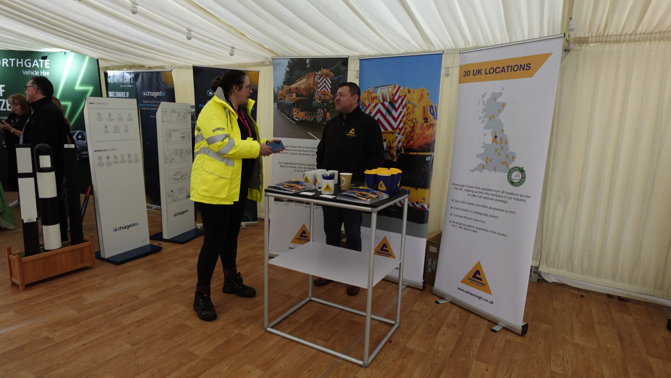 Trade show booth inside a large tent with wooden flooring and white draped ceiling. Two people are conversing—one in a high-visibility jacket and the other in dark branded clothing. The booth features a table with promotional materials including brochures, mugs, and a bowl of candy. Behind them are three banners: one showing a UK map labeled "30 UK Locations," another displaying vehicles with high-visibility markings, and a third partially visible banner related to vehicle hire services.
