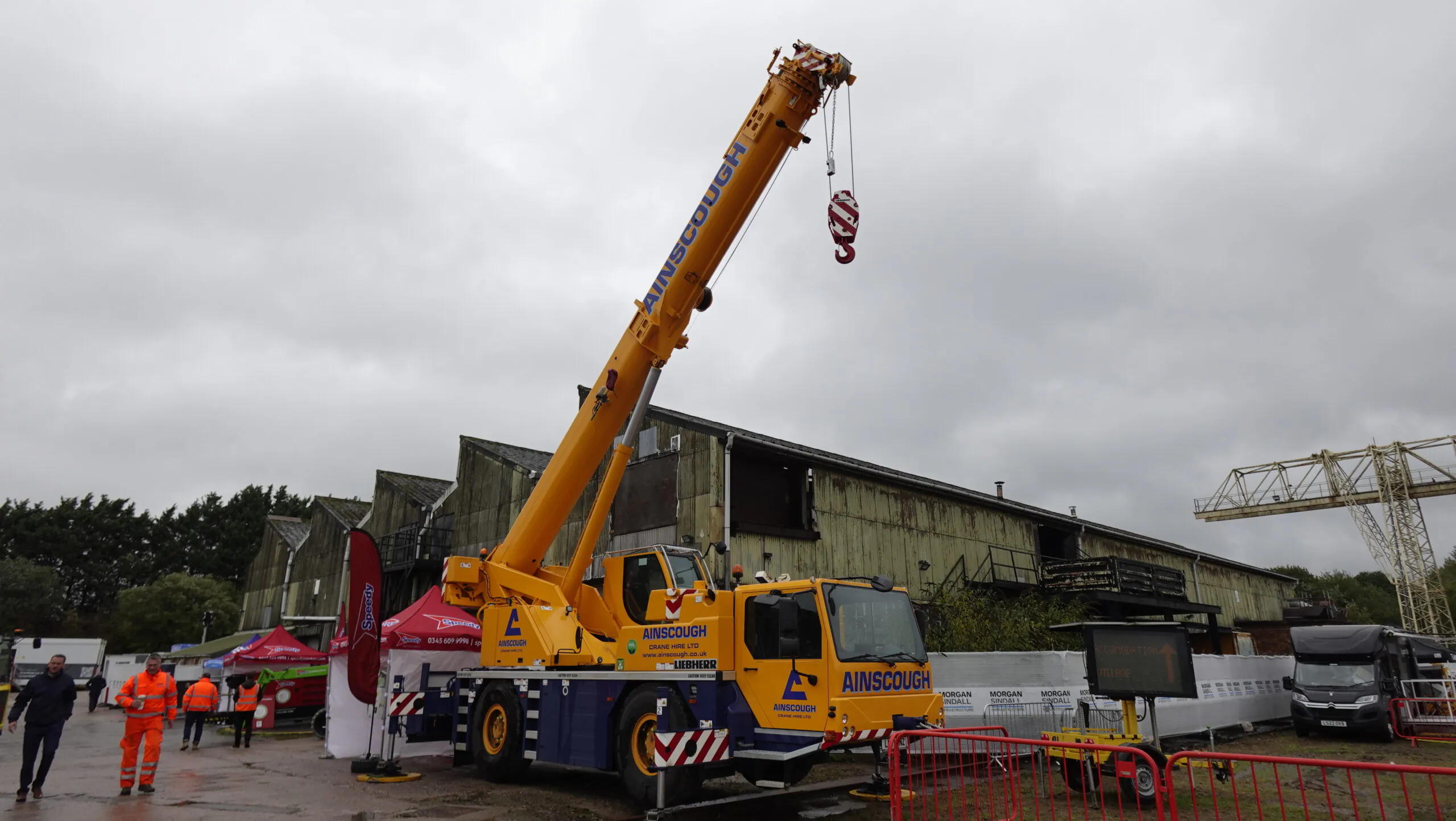 Large yellow crane from Ainscough Crane Hire Ltd parked outdoors with people in high-visibility clothing nearby.