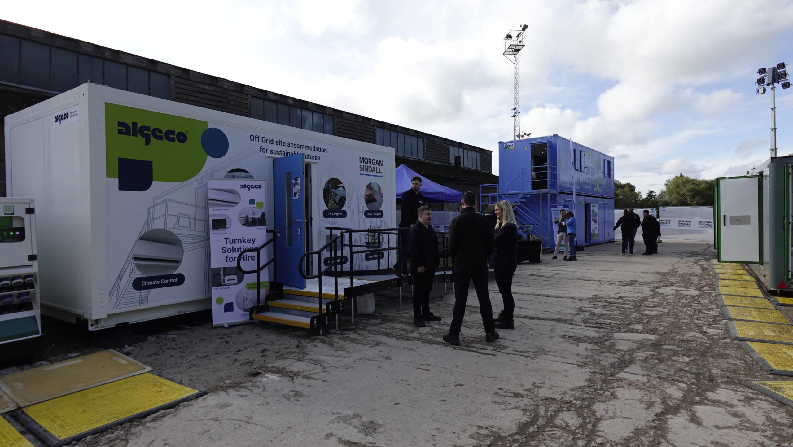 Outdoor setup with white and blue portable buildings labeled "algeco" and "Morgan Sindall"; people are gathered and conversing.