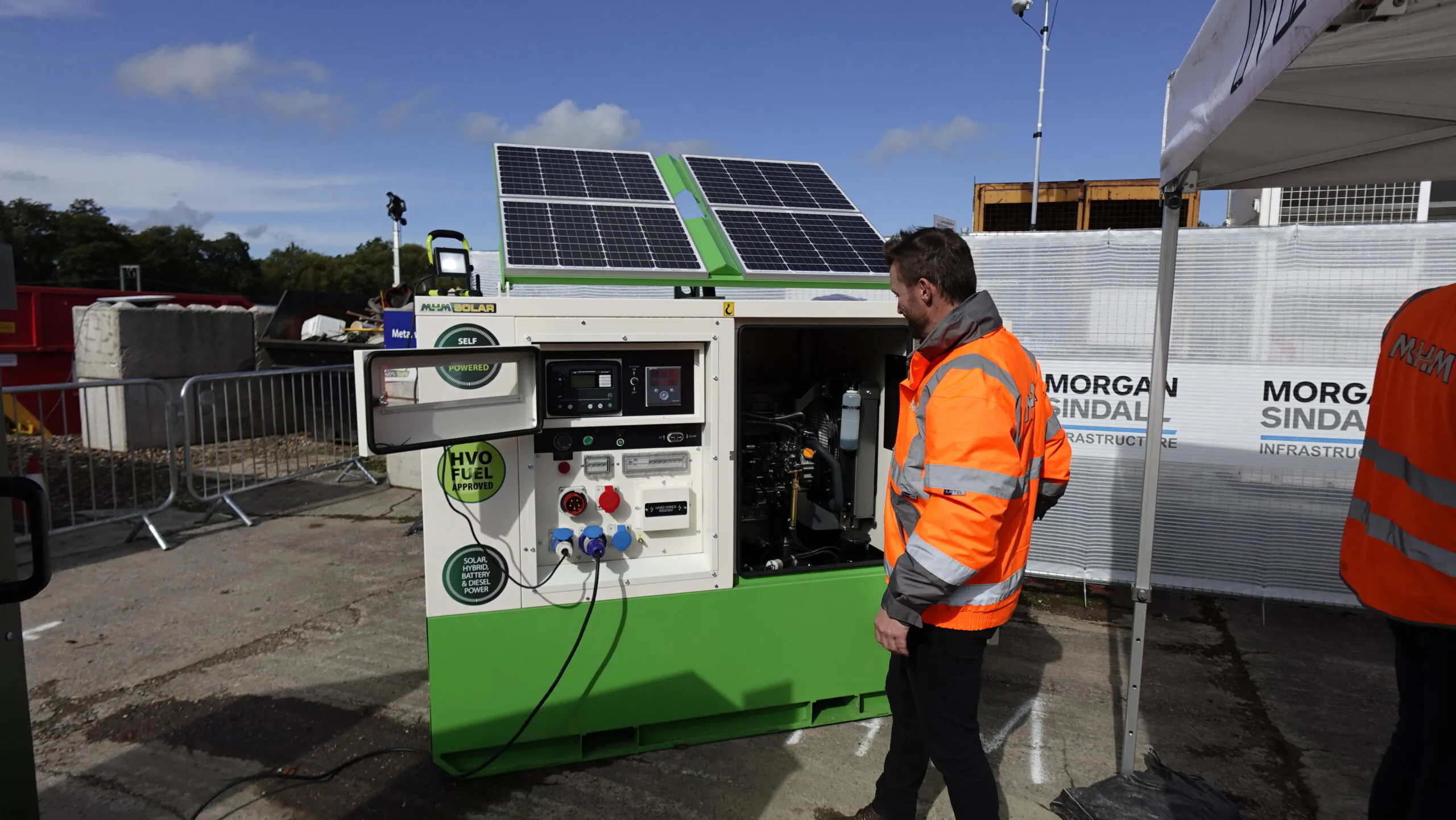 Person in orange high-visibility jacket inspecting machinery with solar panels labeled "HVO Fuel."