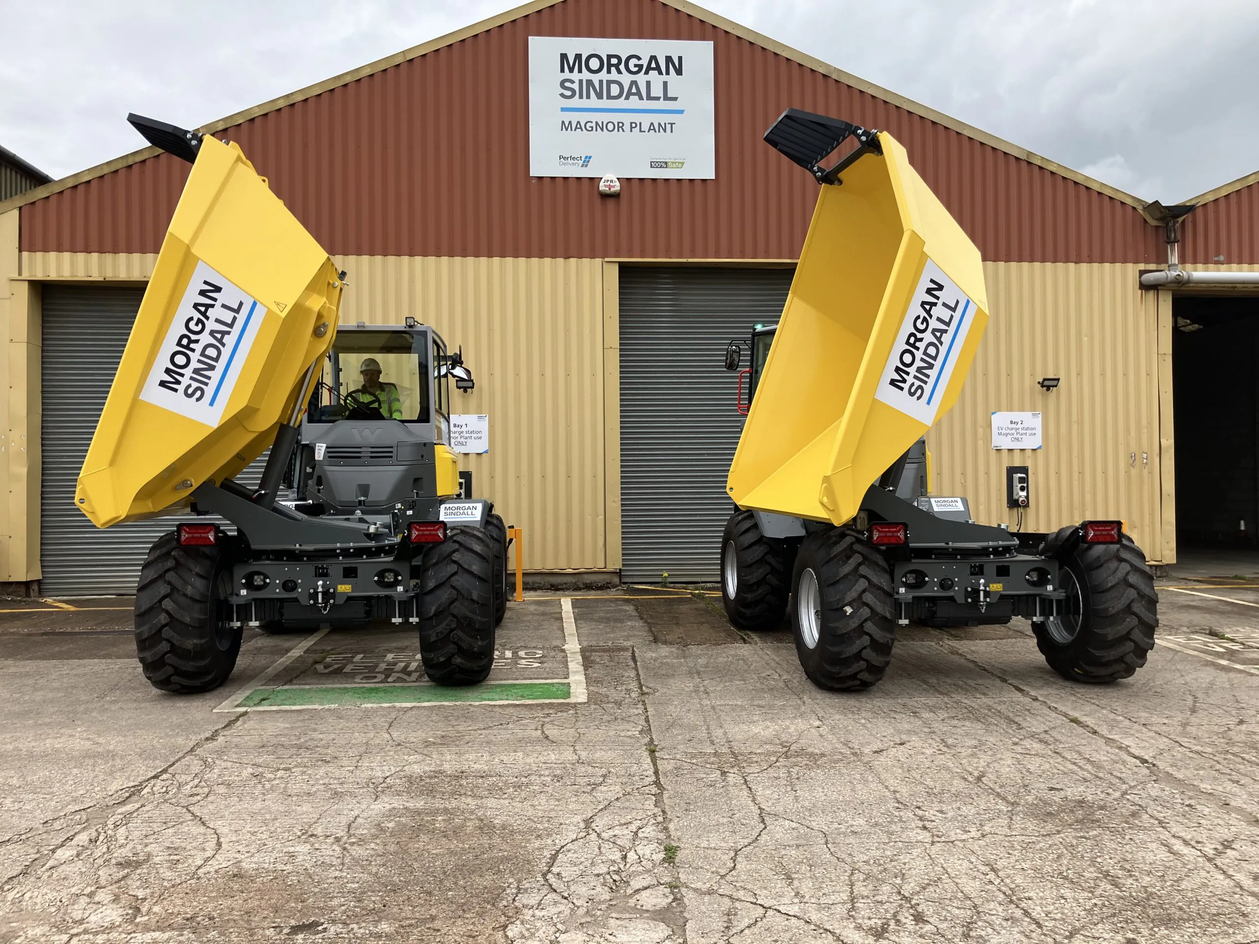 Two large yellow dual-view dump trucks shown from behind, stationed outside the Morgan Sindall Magnor Plant building.