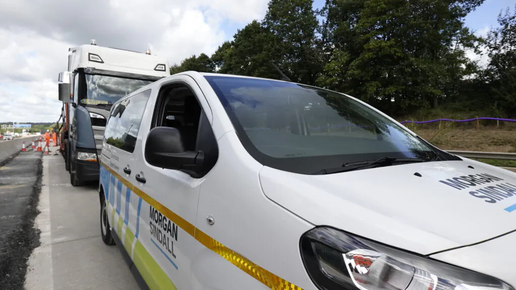 White Morgan Sindall electric minibus parked on the roadside next to a large truck.