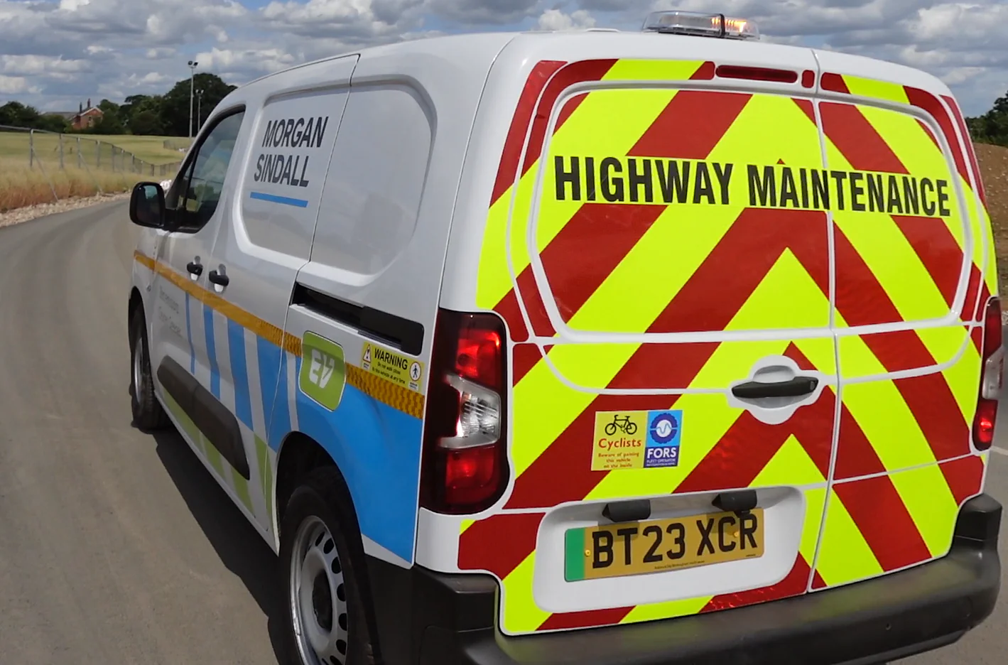 Rear view of a white Morgan Sindall van marked for highway maintenance, driving on an open road under a partly cloudy sky.