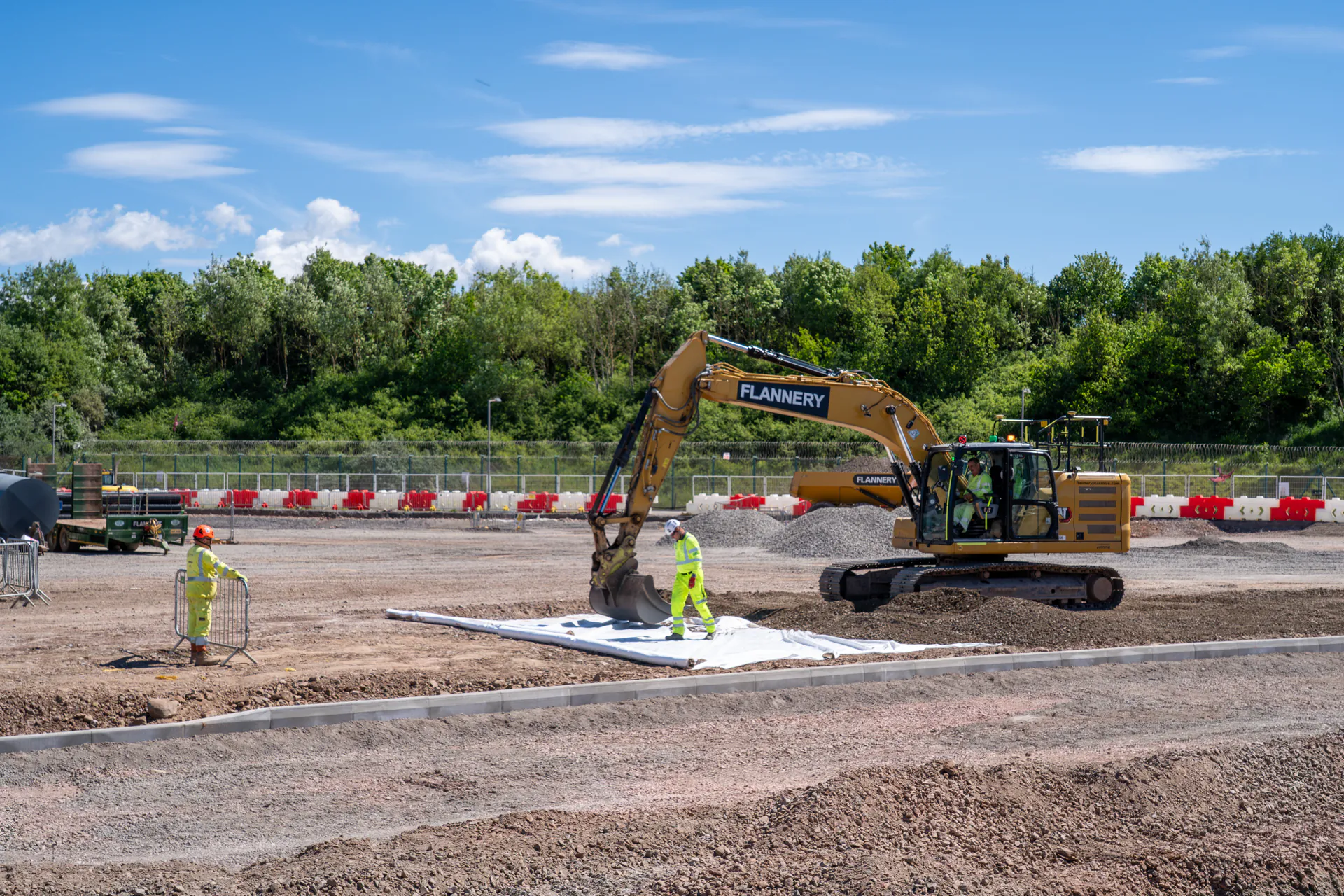 Two construction workers at a site; one operates an excavator while the other stands nearby with equipment, surrounded by red safety barriers.