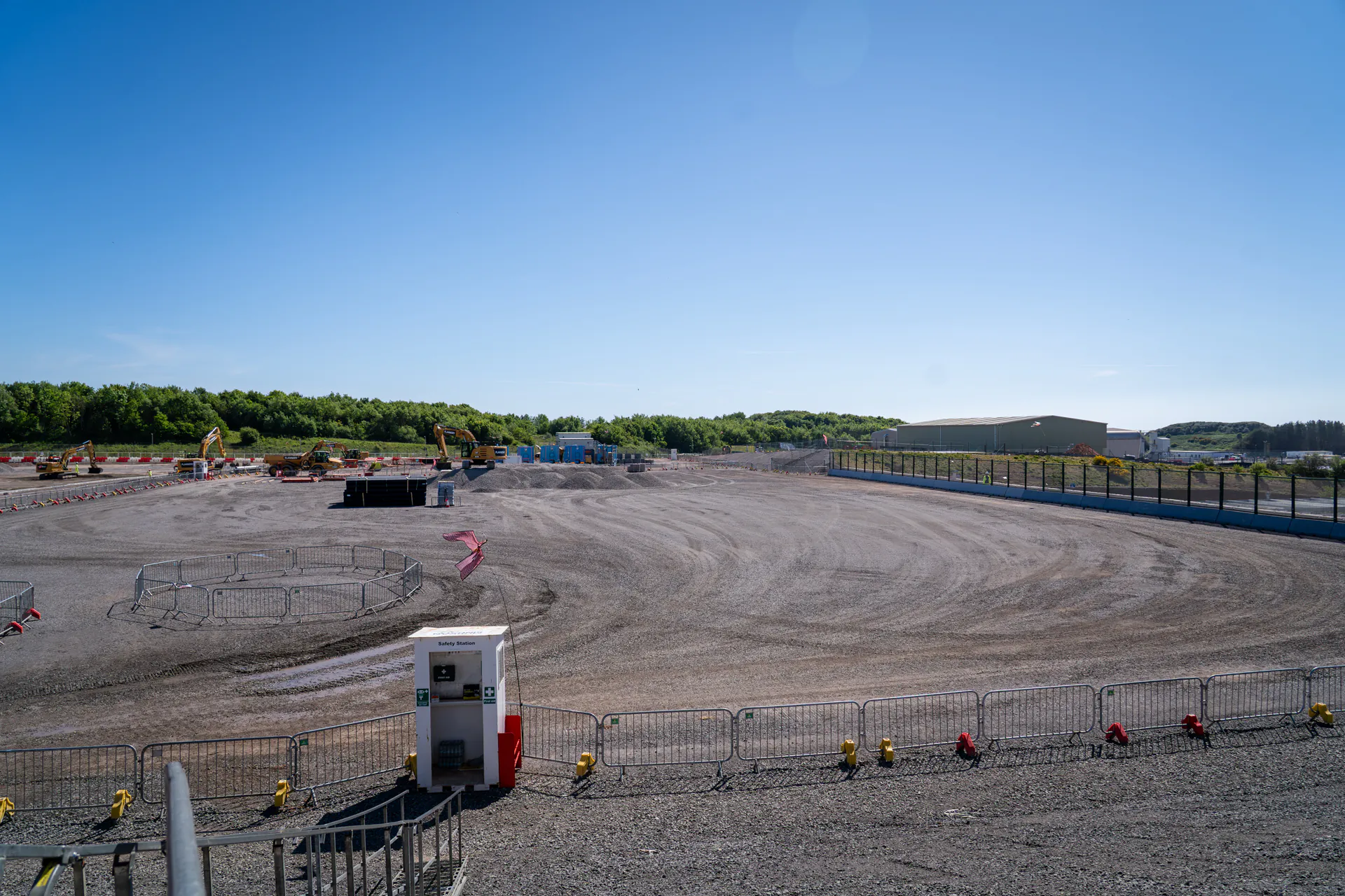 Wide view of a gravel-covered construction site with machinery and metal barriers in the background.
