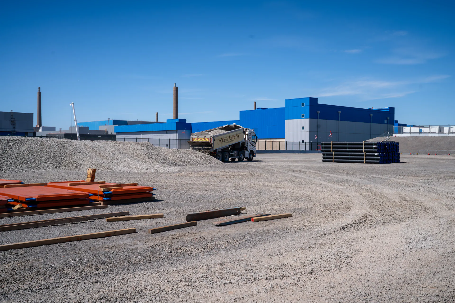Wide view of a construction site with gravel ground, machinery, and fencing under cloudy skies.