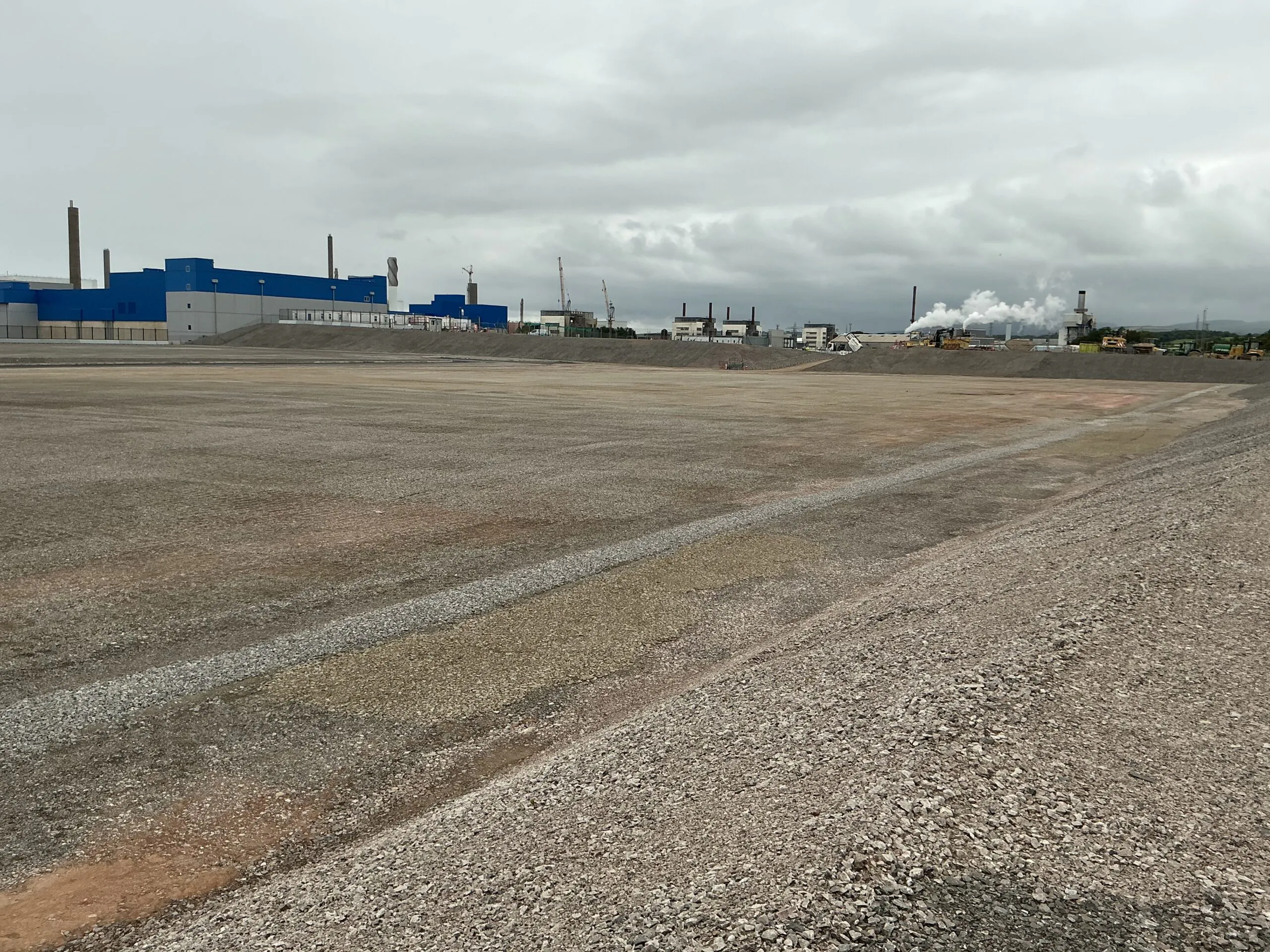 Large empty gravel lot with industrial buildings and smokestacks in the background under an overcast sky.