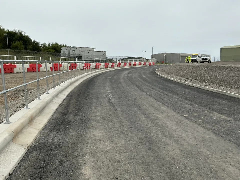 Large empty gravel lot with industrial buildings and smokestacks in the background under an overcast sky.