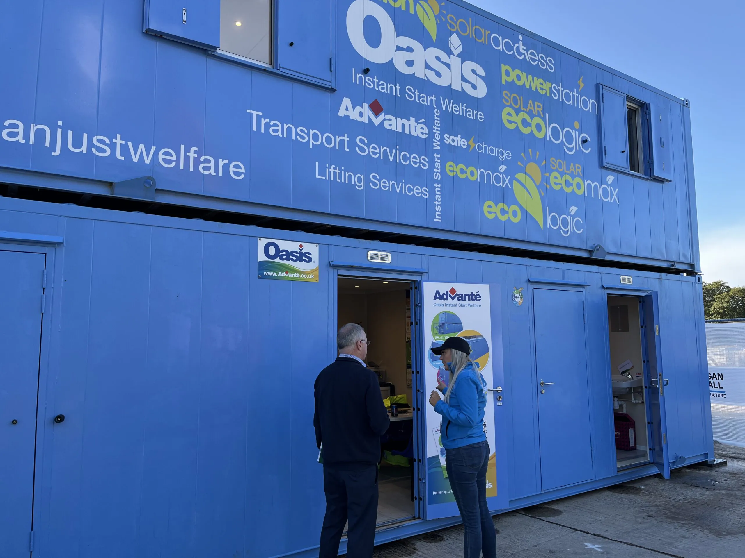 Two people conversing outside near blue portable buildings labeled "Oasis" offering services like transport and lifting by Advanté Ltd.