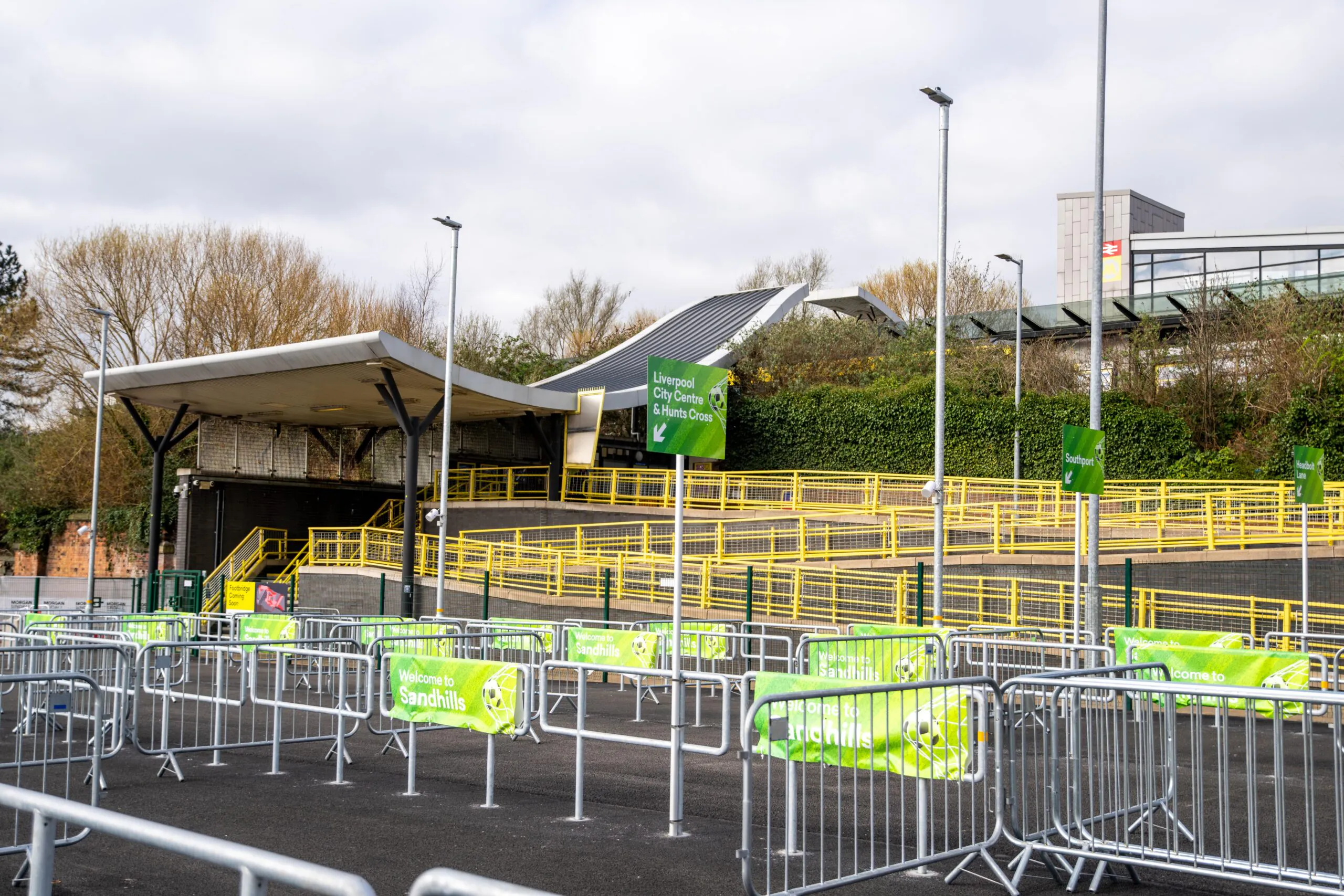 Outdoor area at a Merseyrail station featuring metal barriers arranged for crowd control or queuing, with green directional signs reading "Liverpool City Centre via Kirkdale & Orrell" and "Merseyrail Sandhills." A covered structure with yellow railings is visible in the background, along with trees and a modern building.