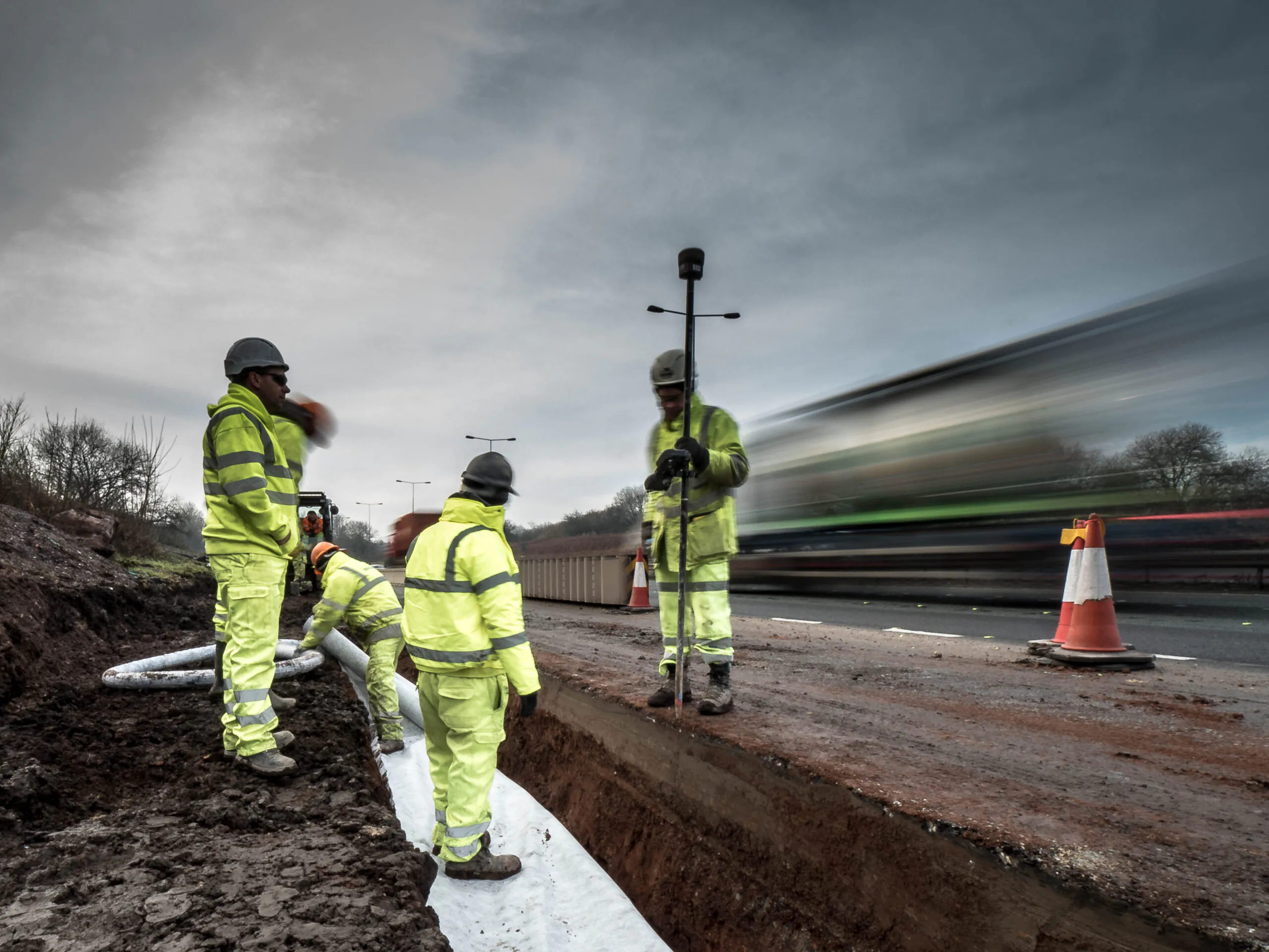 Workers in high-visibility clothing install signage and barriers on a highway under cloudy skies.