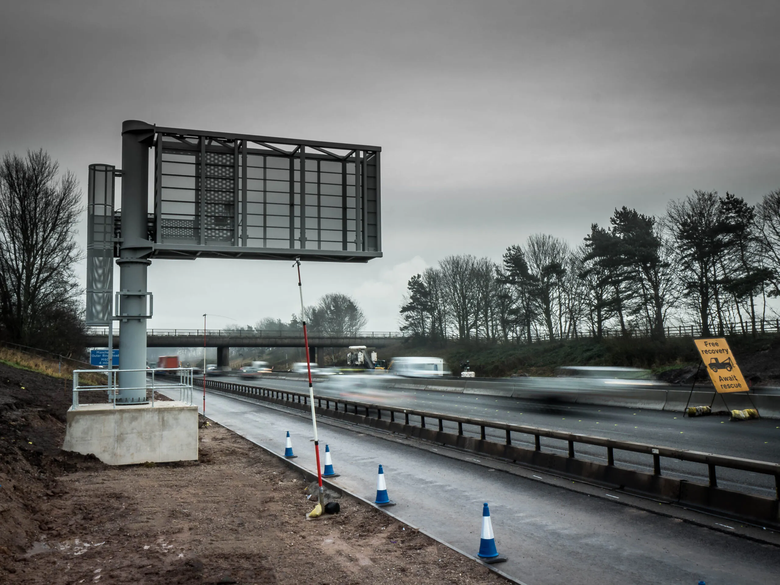 Installation of a large overhead electronic sign structure on a highway under overcast skies, with traffic cones and a yellow sign reading "Free recovery ahead."