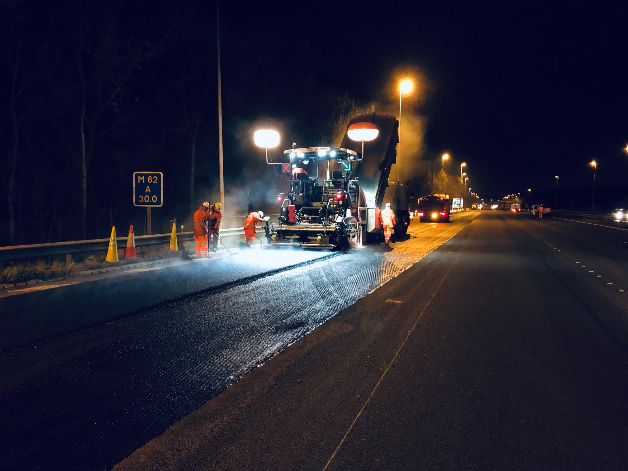 Nighttime roadwork scene with workers resurfacing asphalt on a highway using heavy machinery illuminated by bright lights.