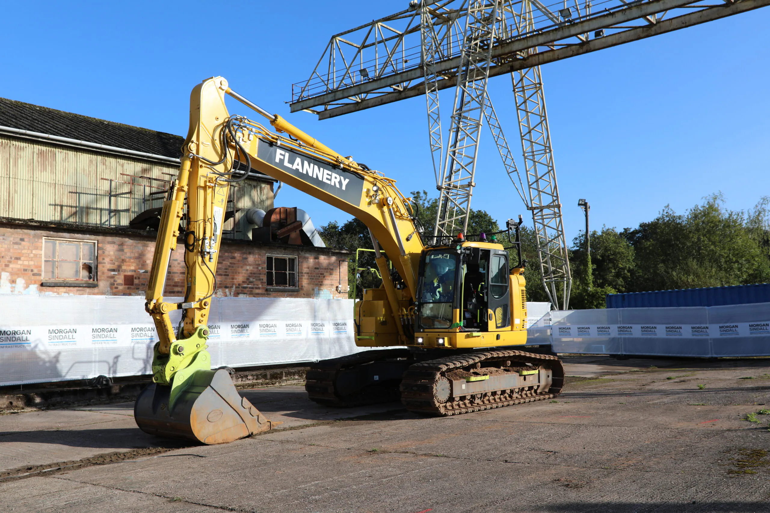 Yellow excavator labeled "FLANNERY" parked outdoors near industrial buildings and scaffolding.