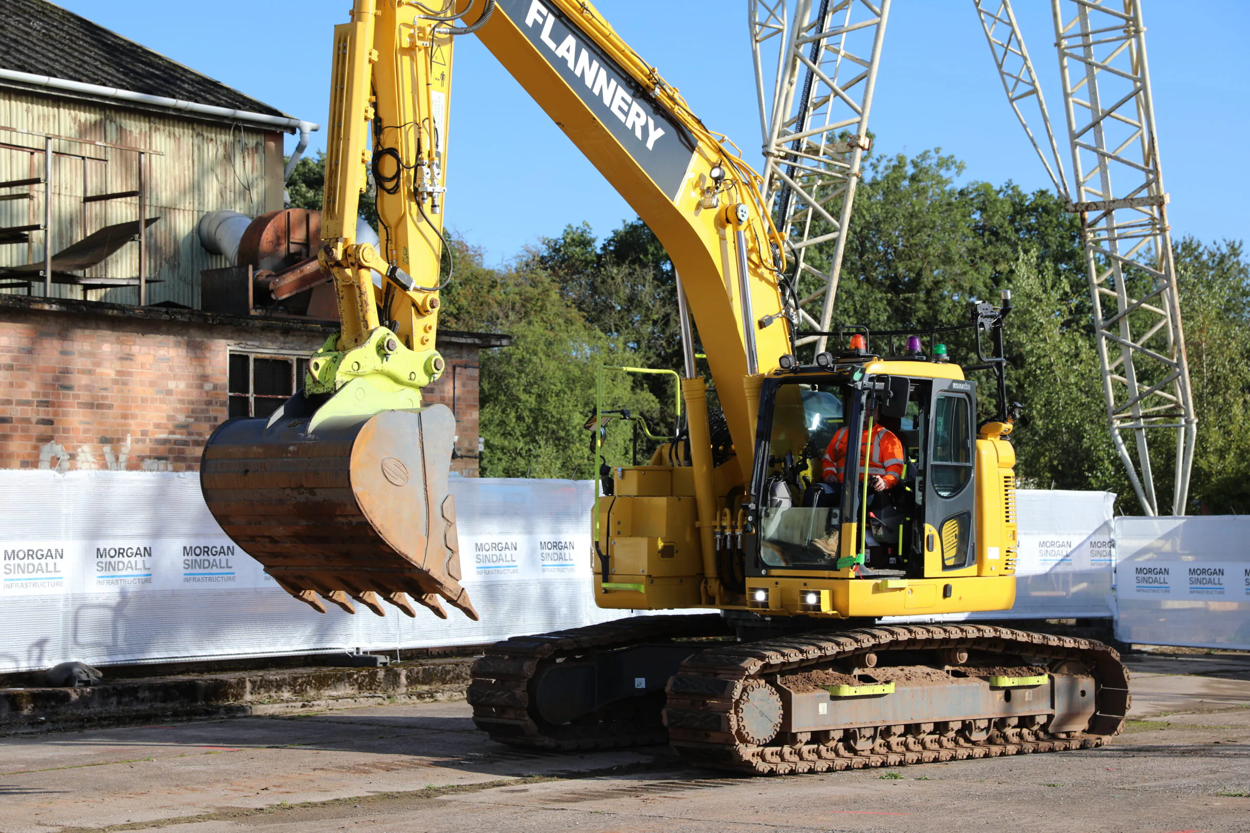 Alternate view of the yellow "FLANNERY" excavator parked outdoors near industrial buildings and scaffolding.