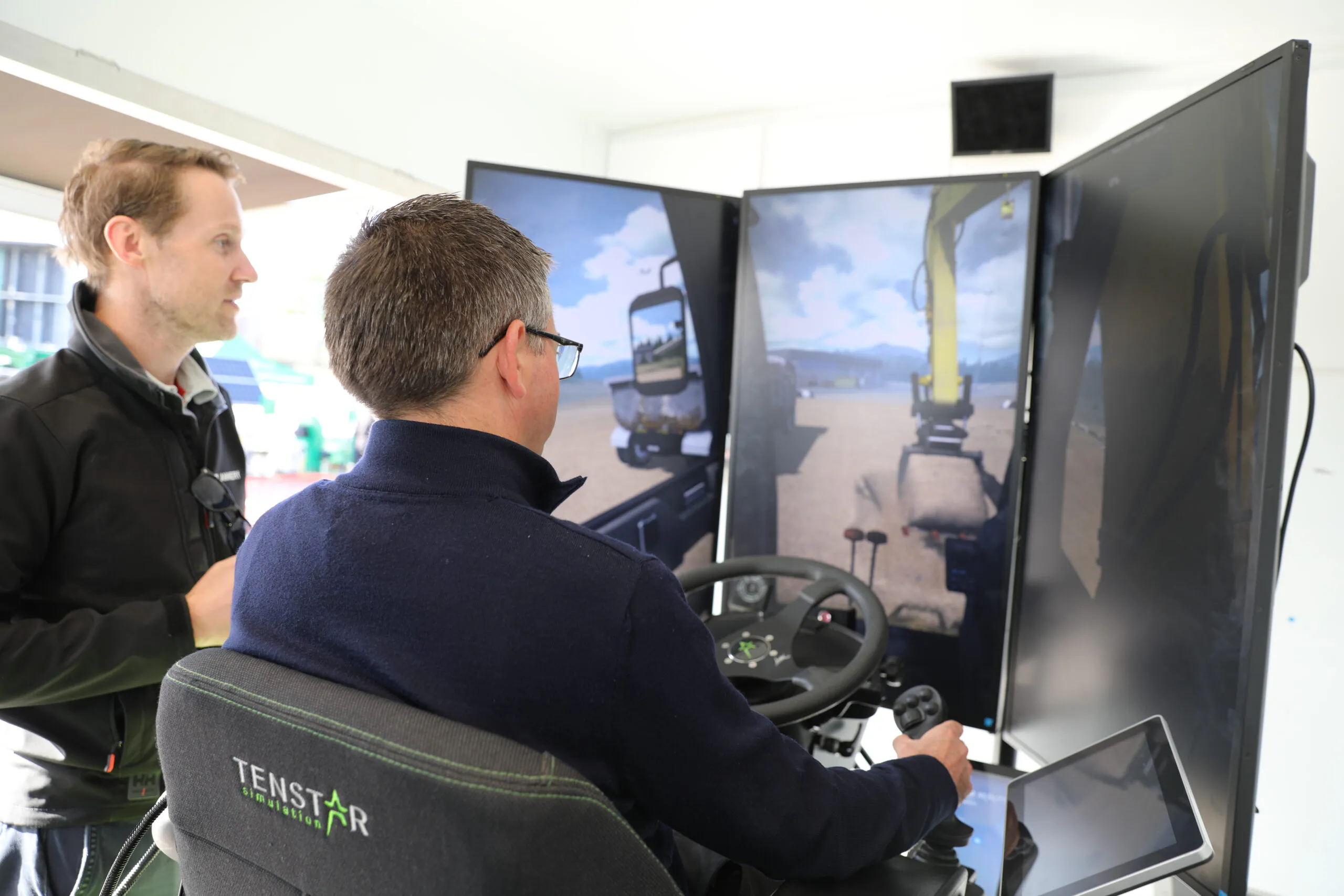 Two individuals interacting inside an equipment simulator setup with multiple screens showing construction machinery controls; Tenstar Simulation AB logo visible.