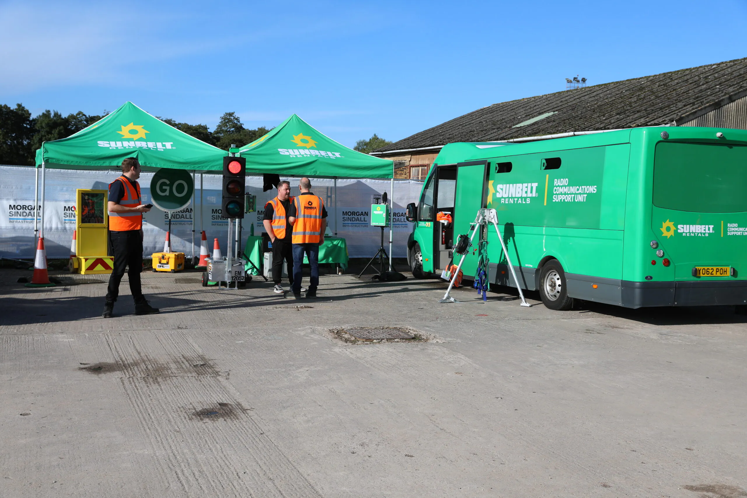 Construction site with two green "Sunbelt Rentals" tents and a green van; people in high-visibility vests are gathered near a traffic light displaying "GO."