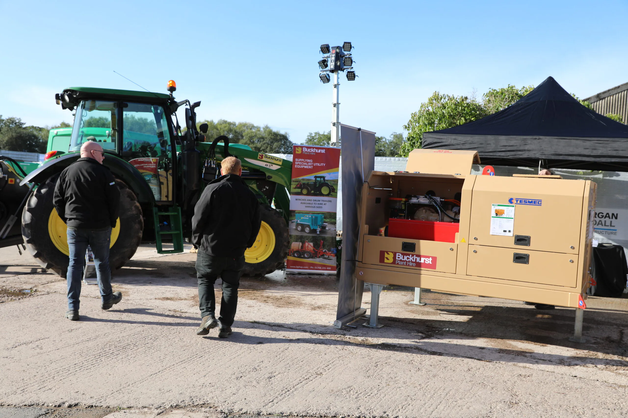 Two men standing beside agricultural machinery and generator setup under tents; Buckhurst Plant Hire Ltd branding visible.