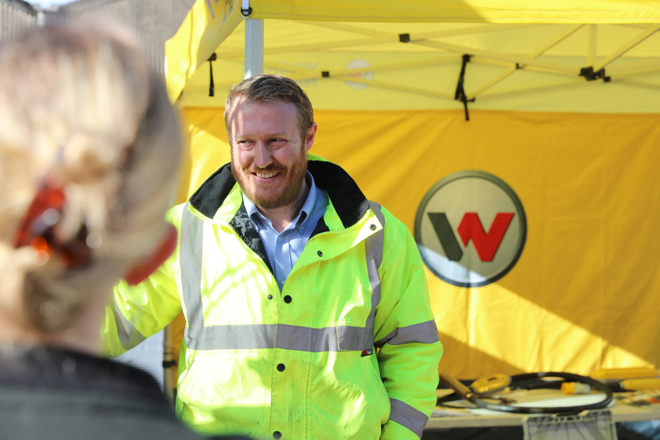 Person in a bright yellow jacket standing under a yellow "Wacker Neuson" tent, appearing to be in conversation.