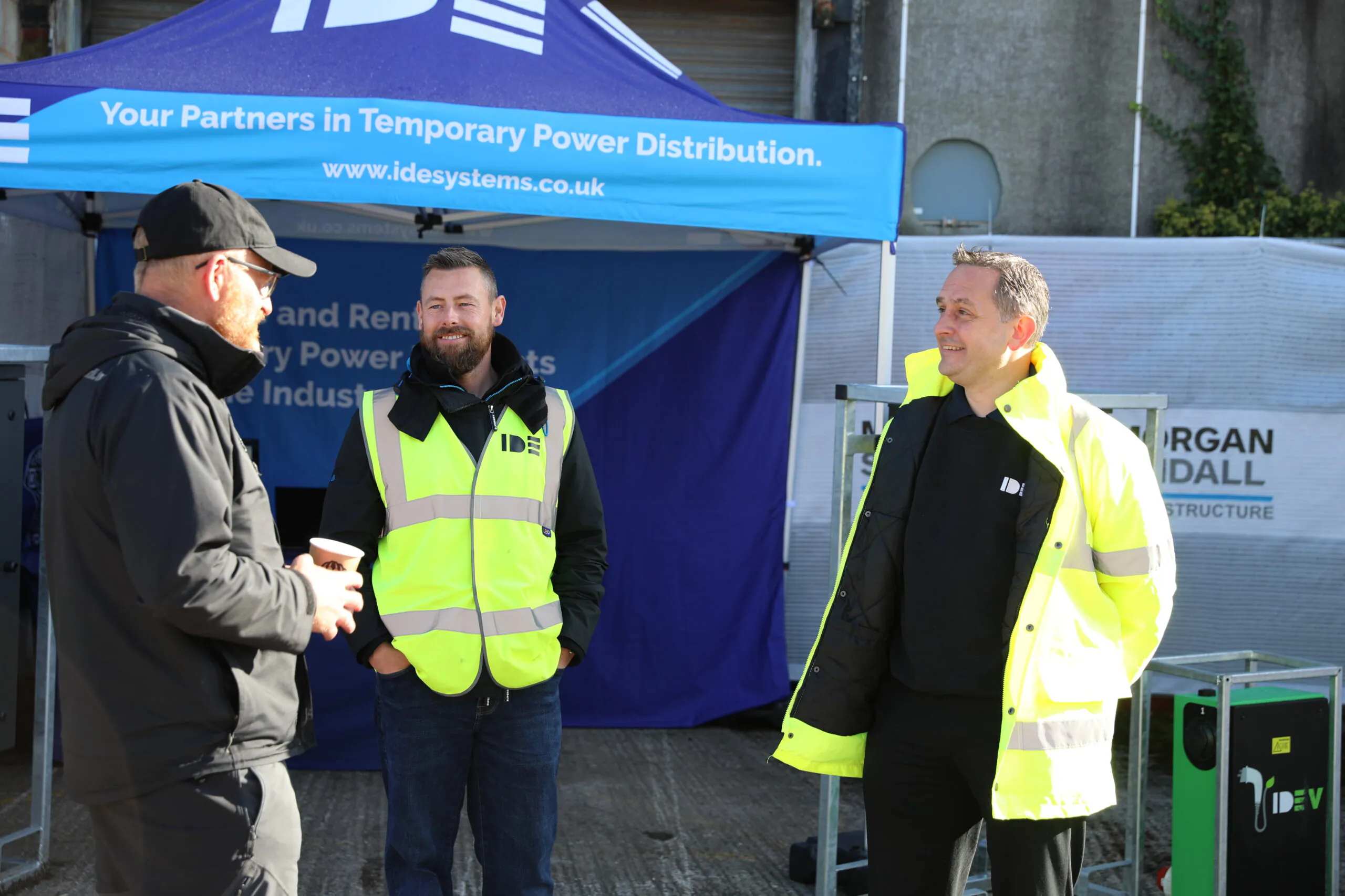 Three individuals talking near an IDE Systems booth under a blue canopy labeled "Your Partners in Temporary Power Distribution."