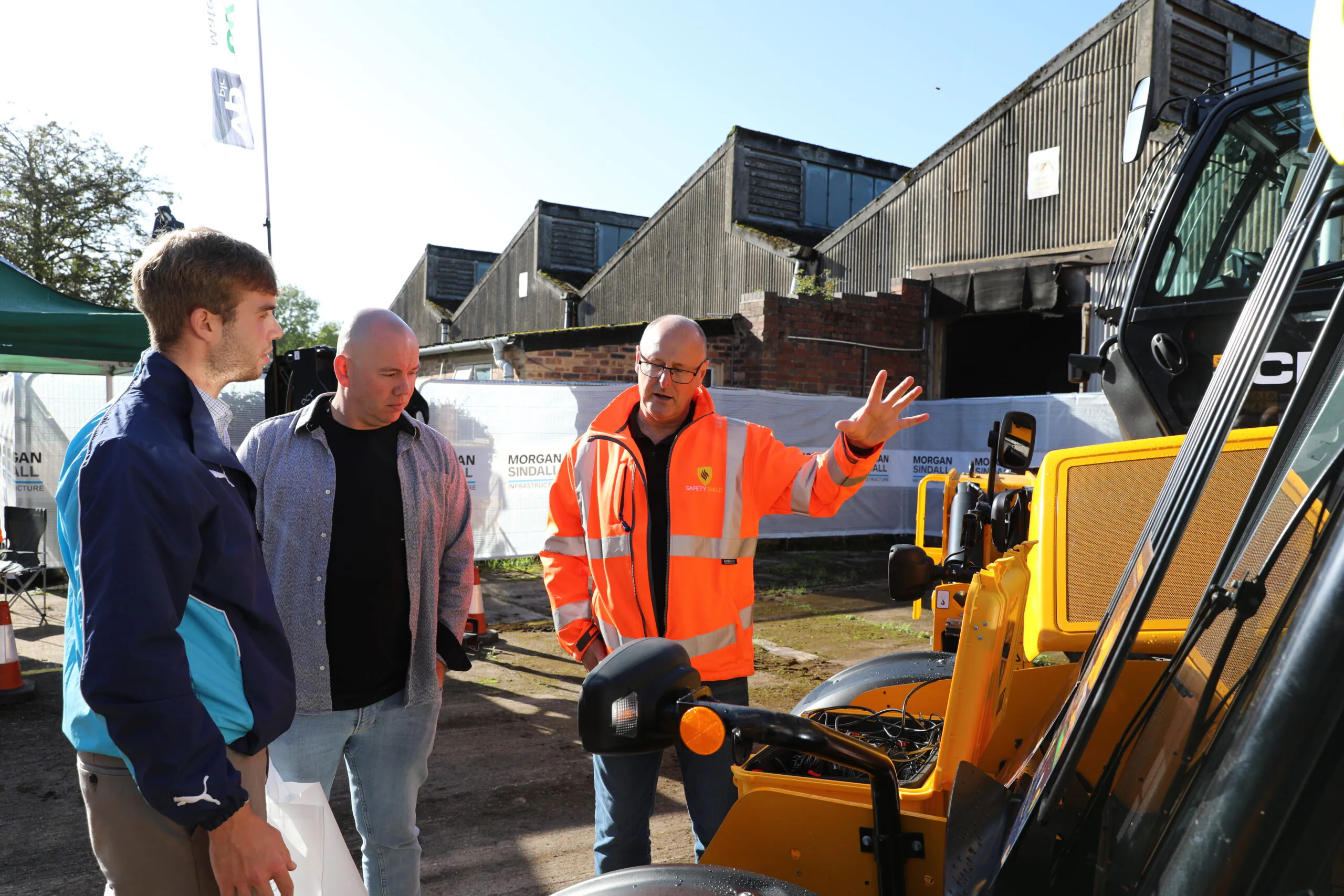 Three individuals near construction machinery, with one person gesturing as if explaining something to the others.