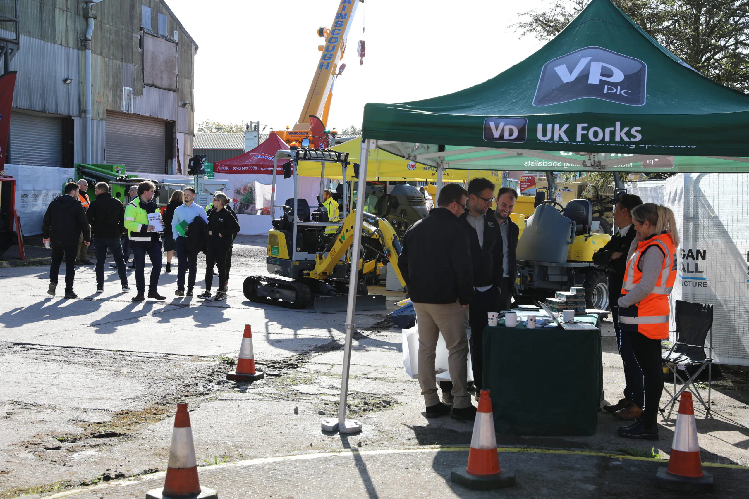 Group of people under a green "VP UK Forks" tent with construction equipment, including a small excavator, visible in the background.
