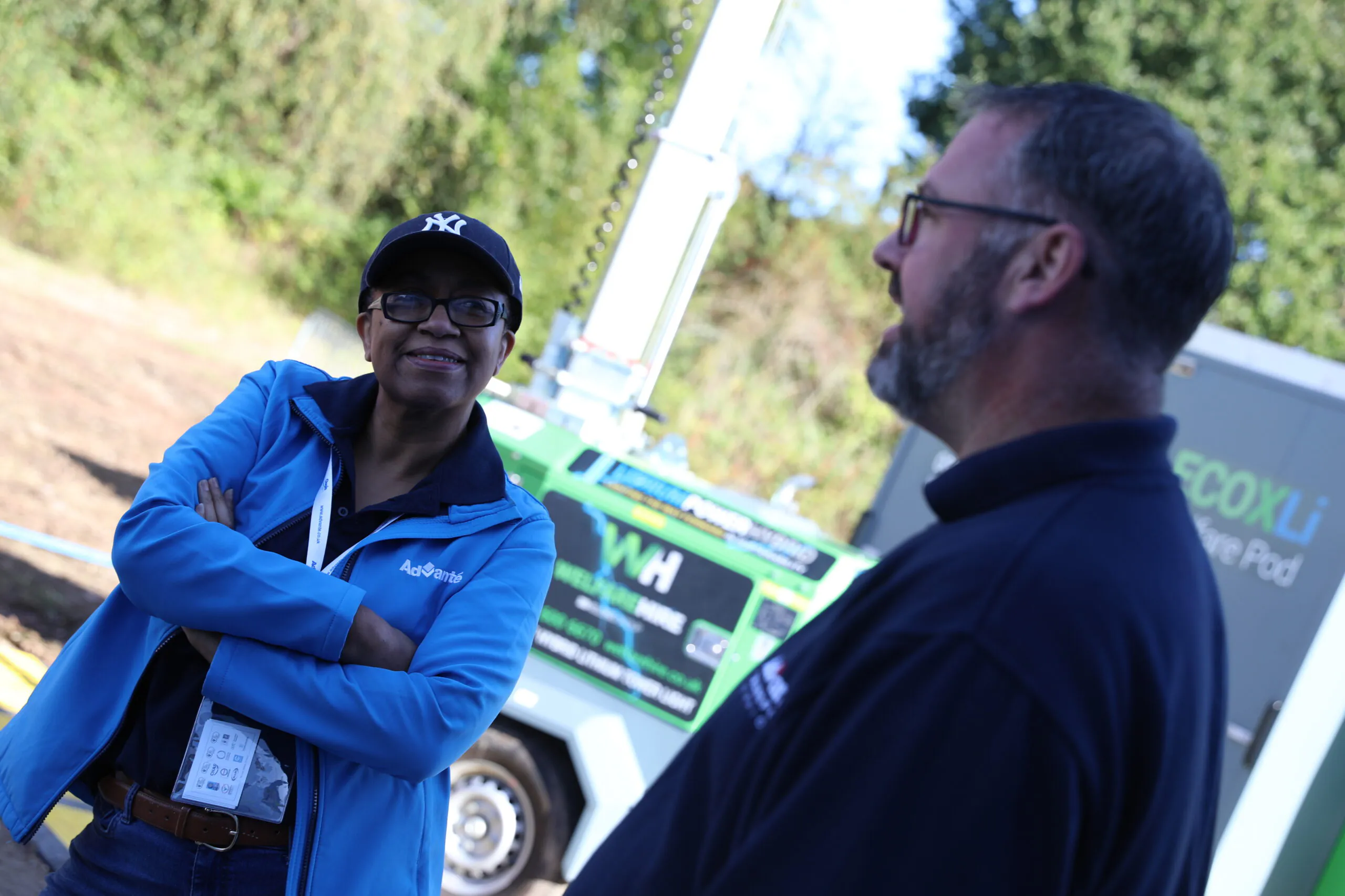 Two individuals conversing outdoors near equipment labeled "WH"; one wears a blue jacket and cap, the other in dark clothing.