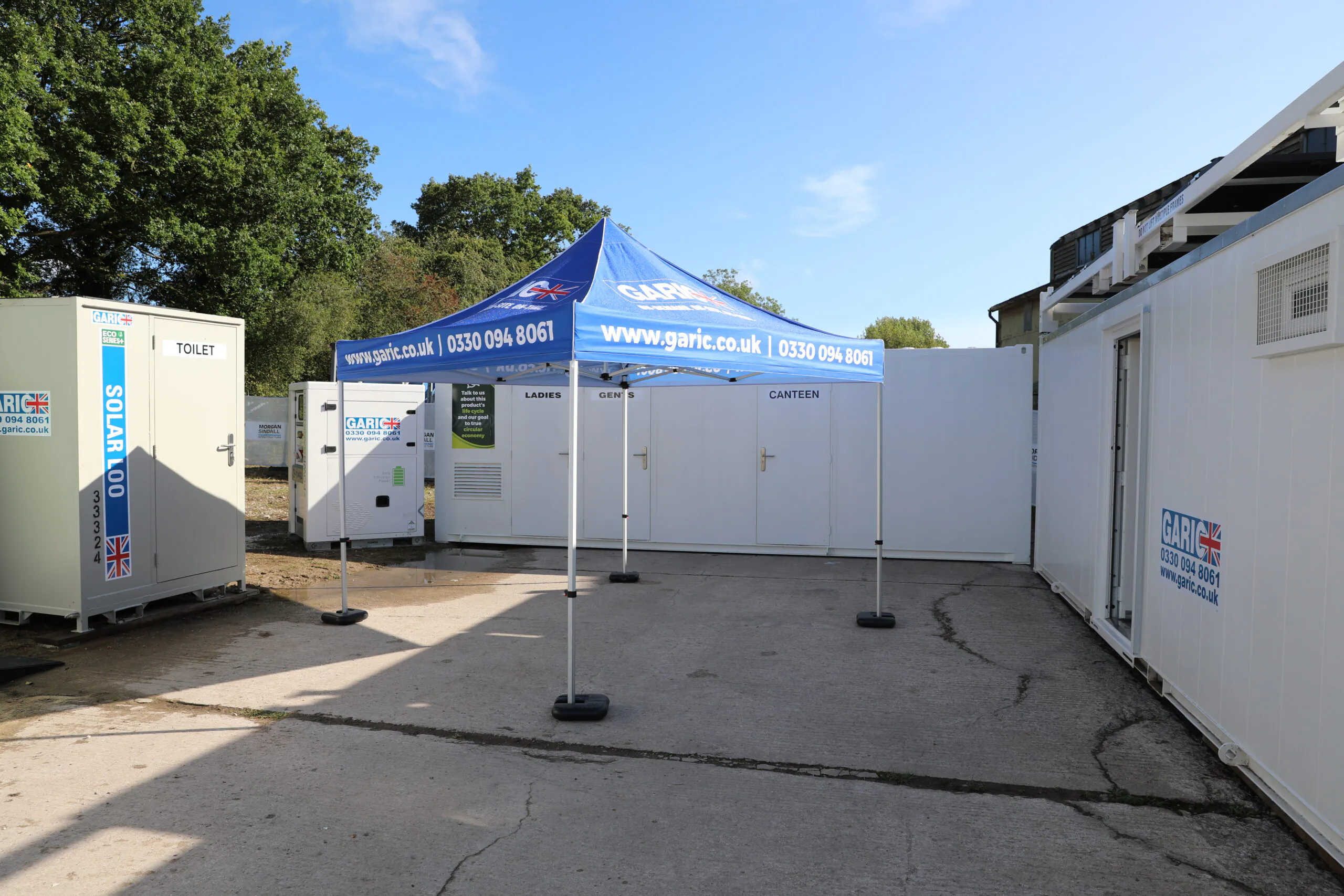 Outdoor setup with portable buildings labeled toilets, canteen, and drying room under blue Garic canopies.